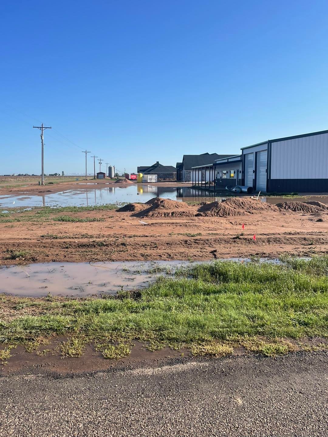 Flooded dirt lot with buildings and power lines under a blue sky.