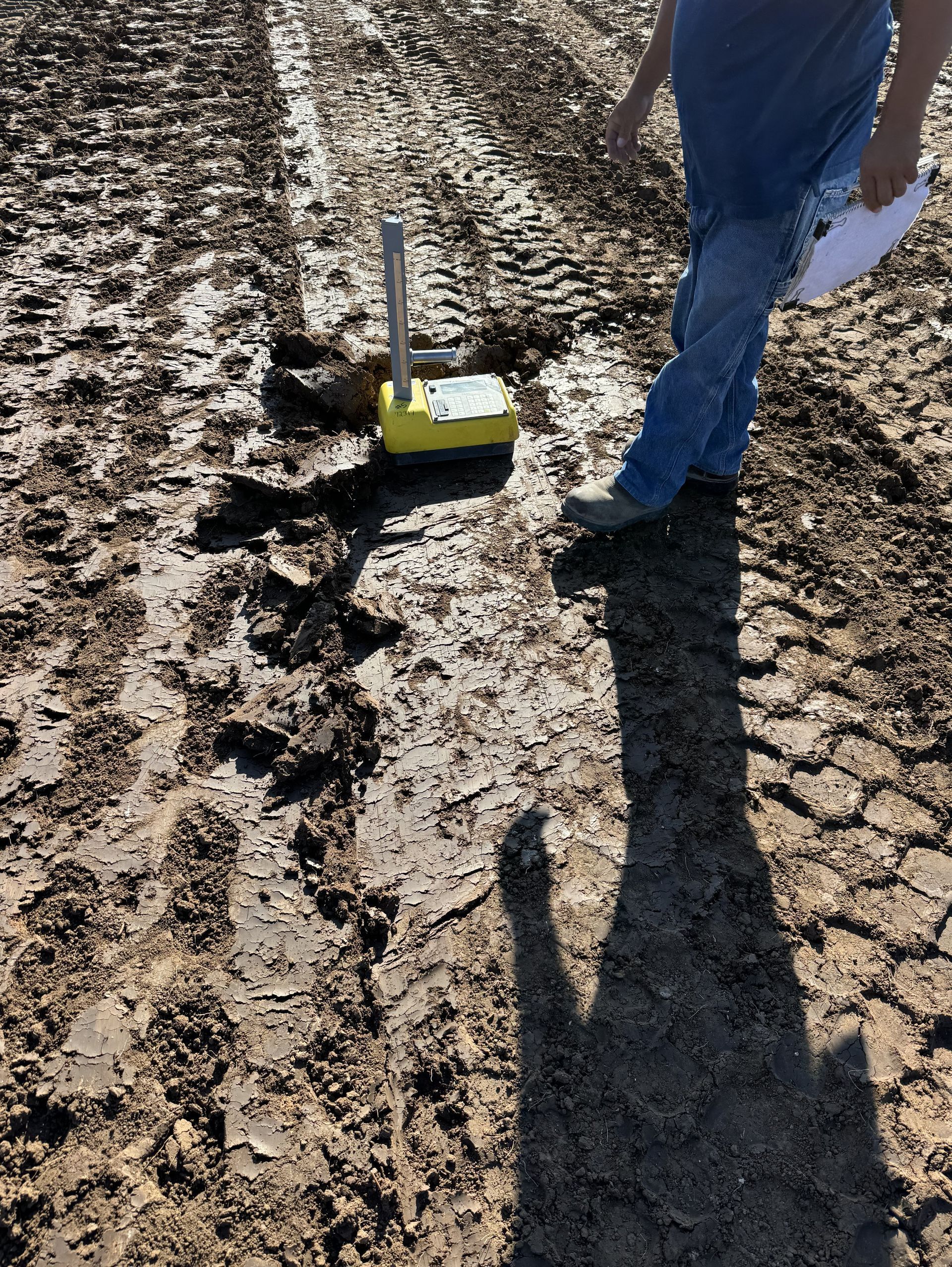 Person stands next to a yellow testing device on muddy ground, with vehicle tracks visible.