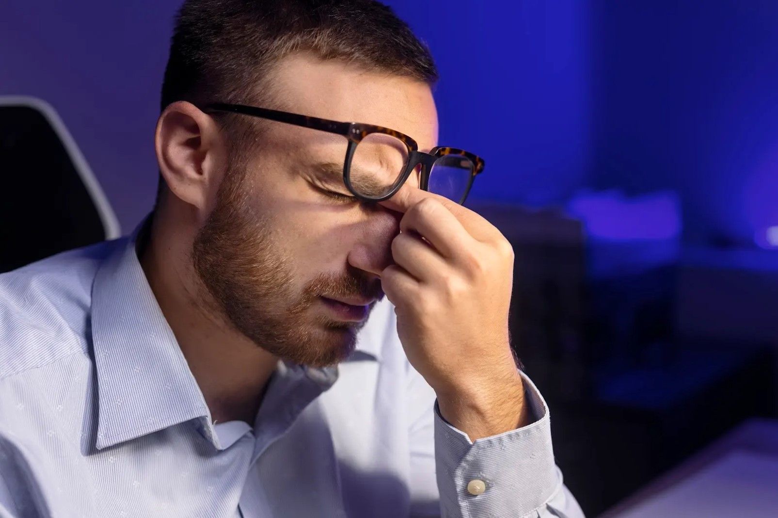 Man wearing glasses presses fingers to bridge of nose, looking tired, in a dimly lit room.