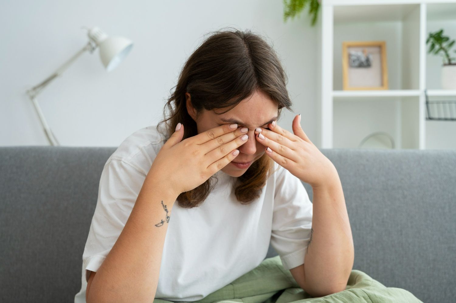 Woman sitting on couch, rubbing eyes with both hands. She wears a white shirt in a living room.