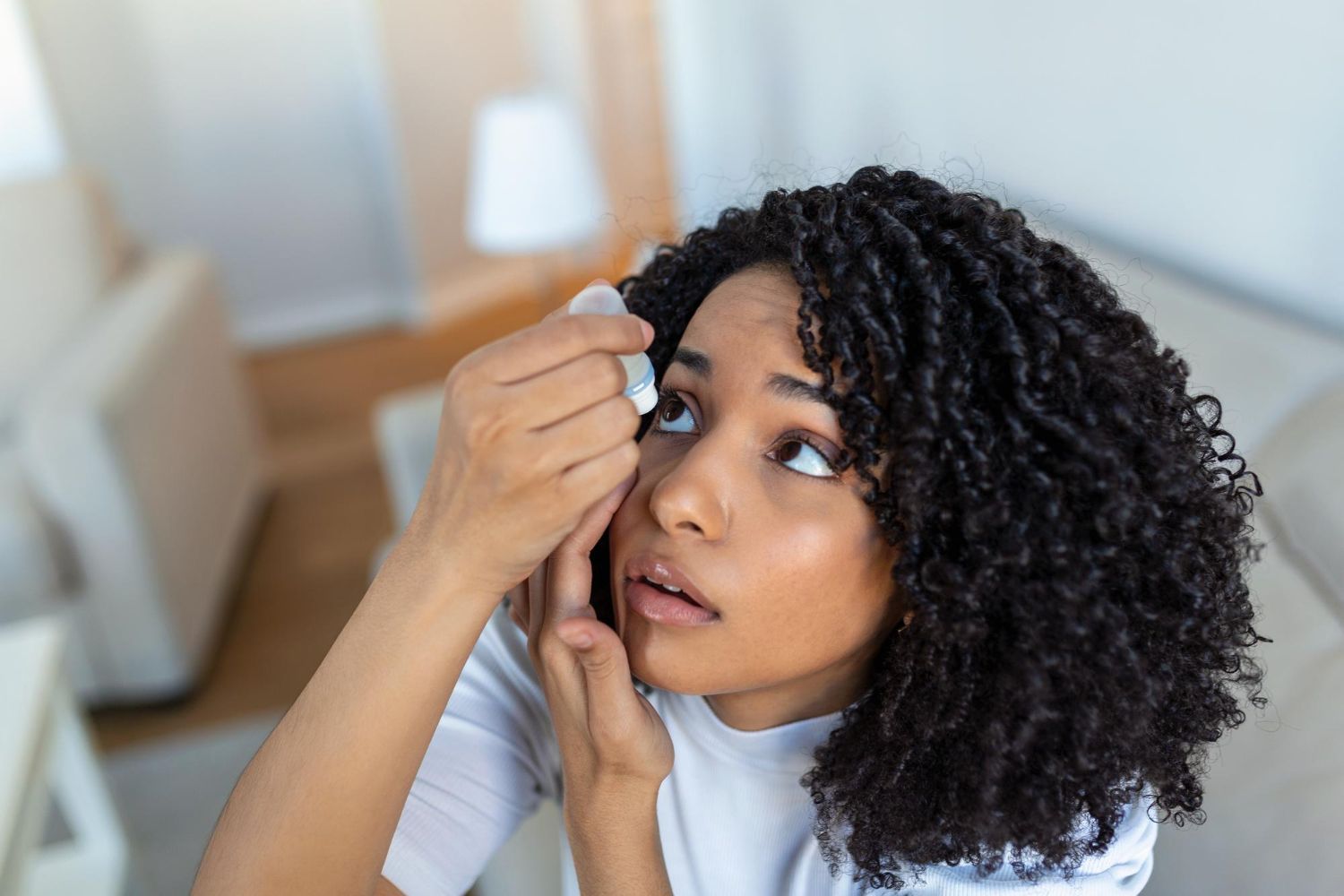 A woman is applying eye drops to her eye while sitting on a couch.