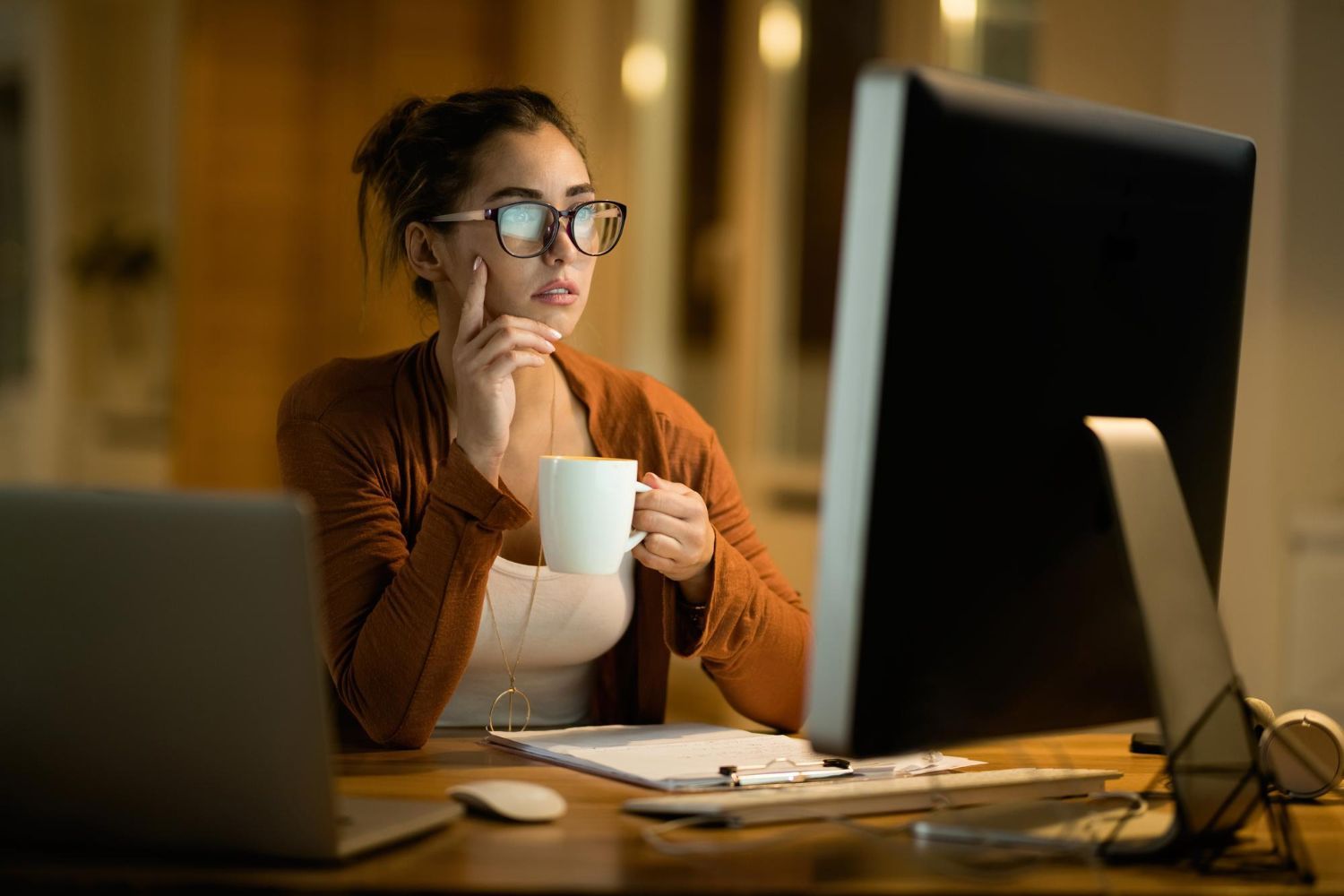 A woman is sitting at a desk drinking coffee and looking at a computer screen.