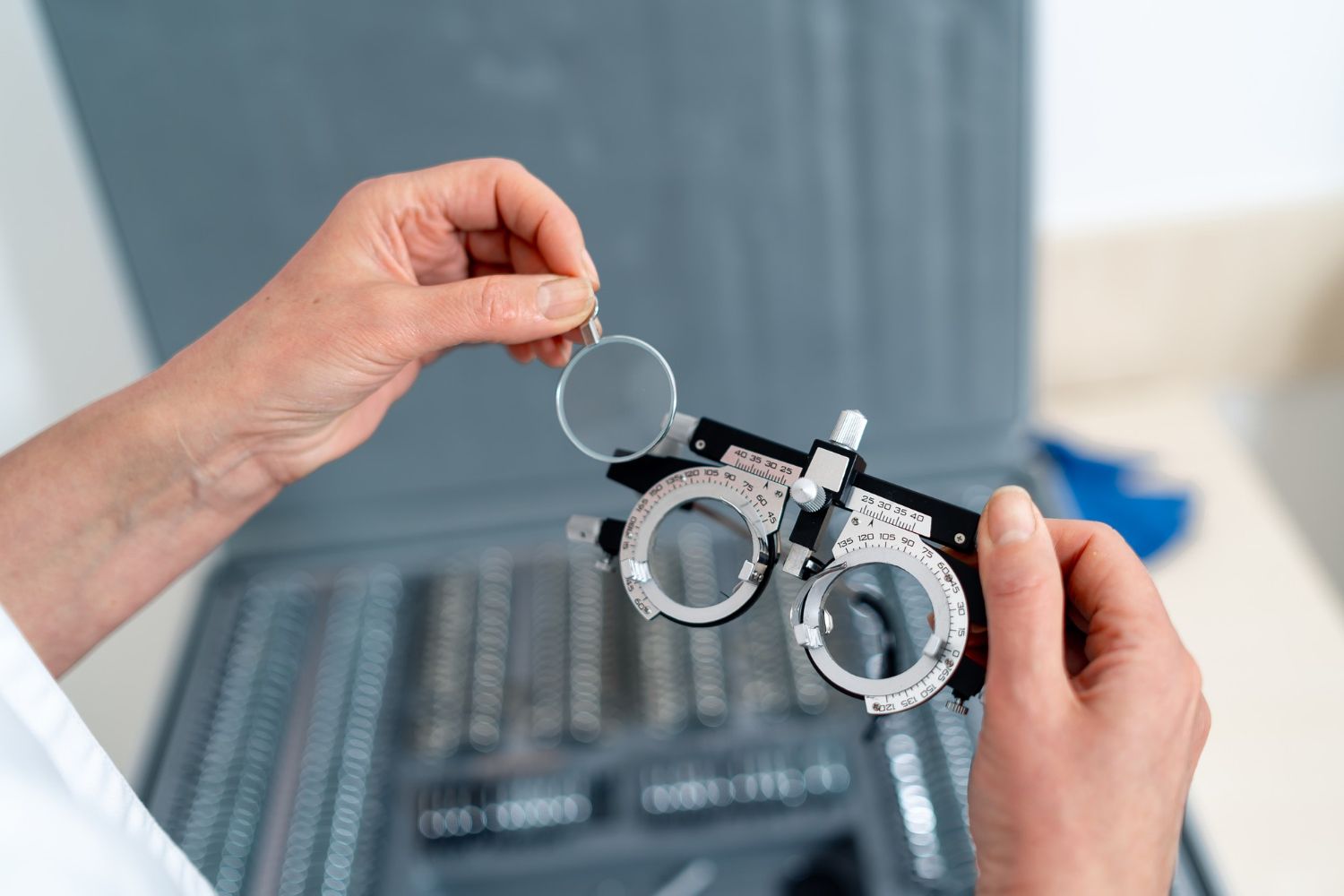 Person holding a trial frame with a lens, near an open case of lenses, in a medical setting.