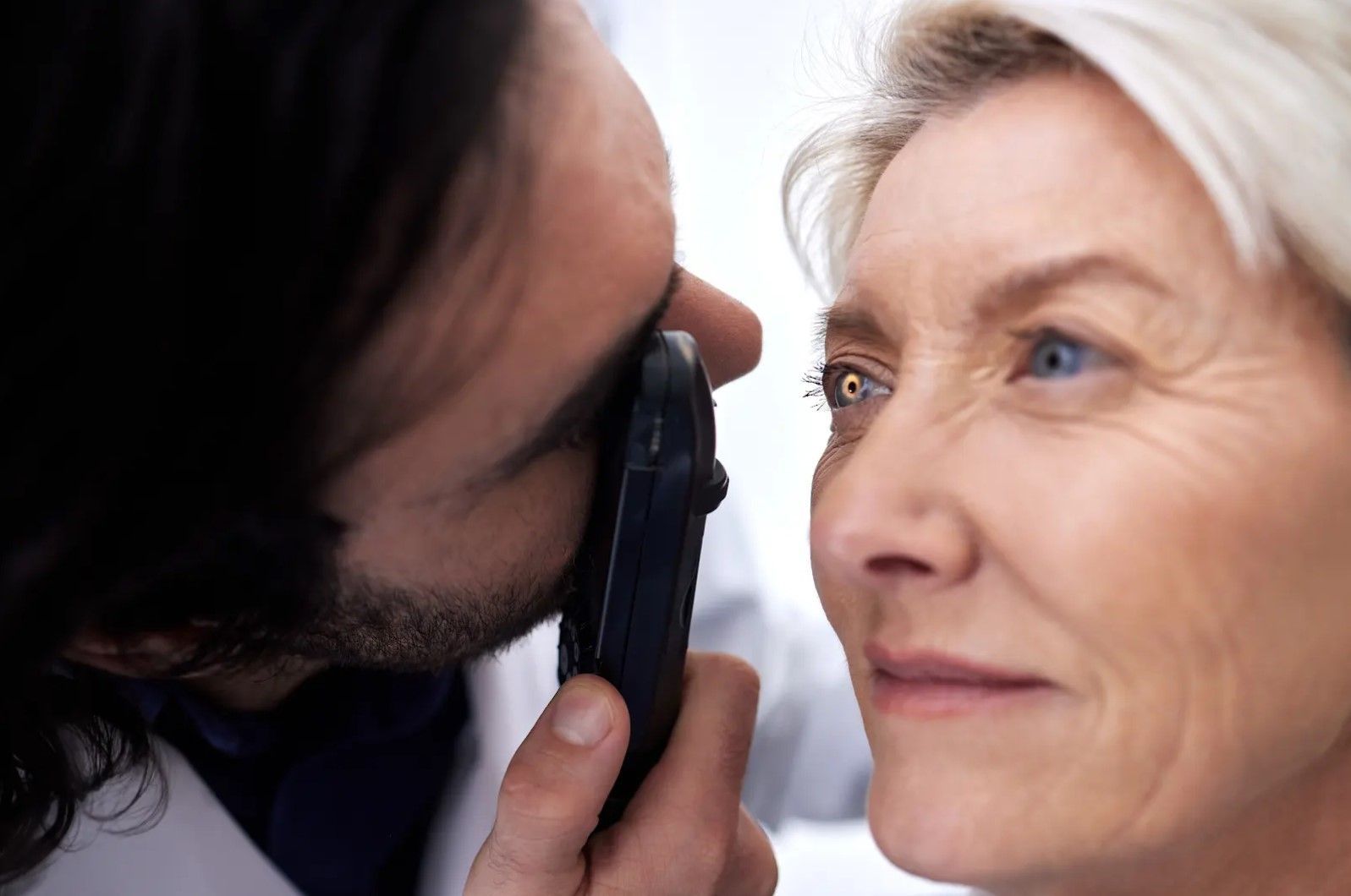 Optometrist examining a senior woman's eye with a handheld ophthalmoscope. Both are indoors.