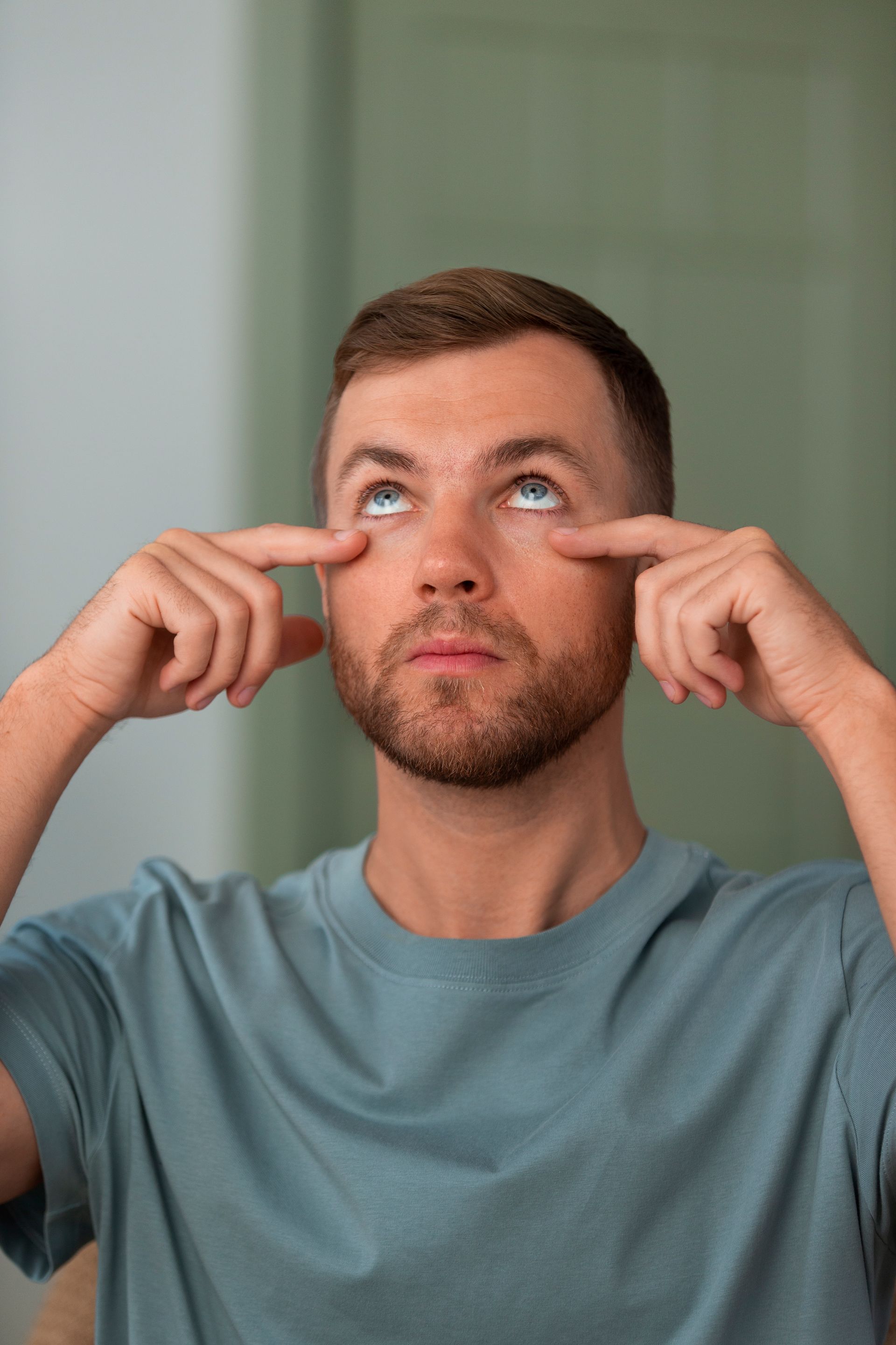 A man with a beard is touching his eyes in front of a mirror.