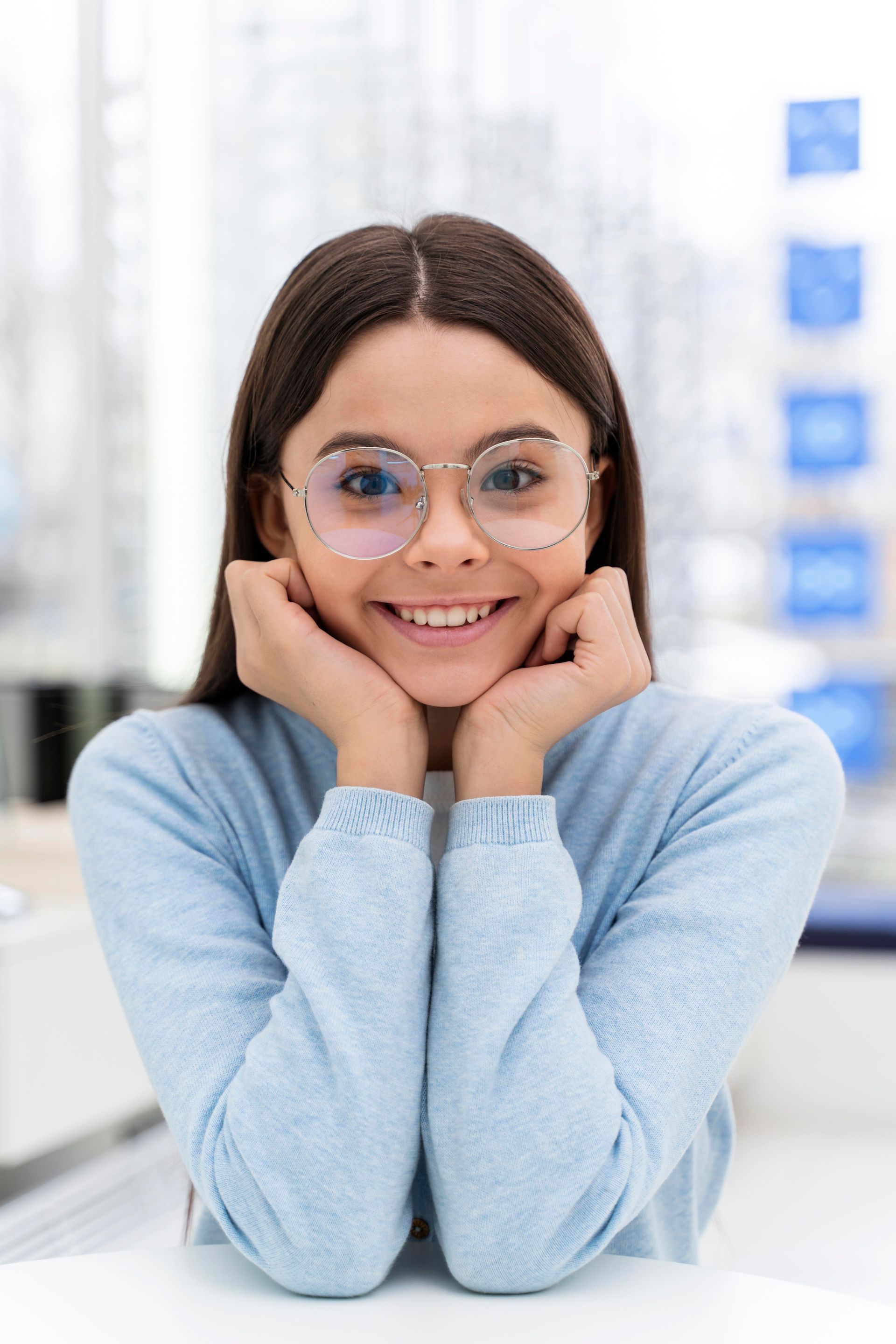 A young woman wearing glasses and a blue sweater is smiling.