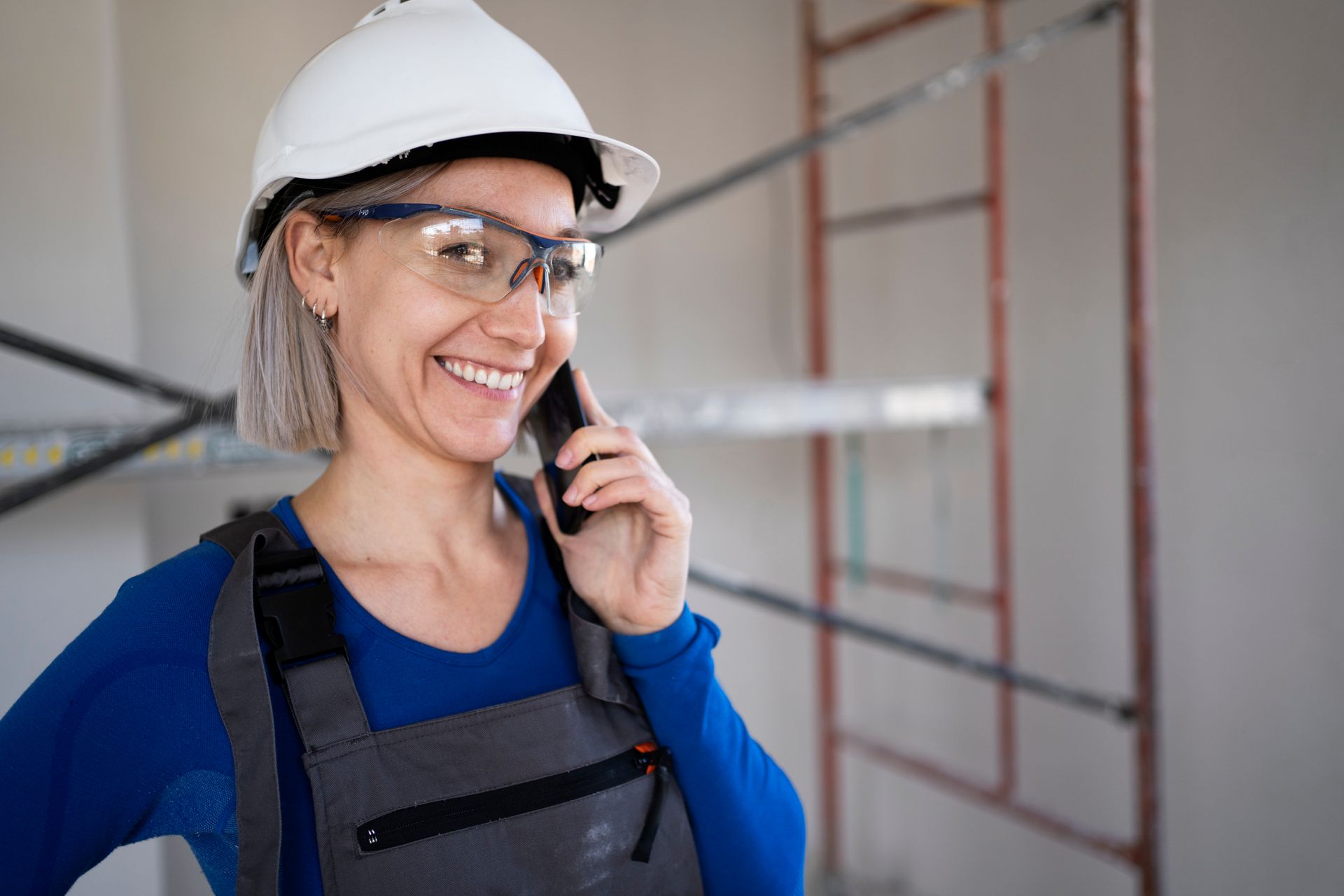 A woman wearing a hard hat and safety glasses is talking on a cell phone.