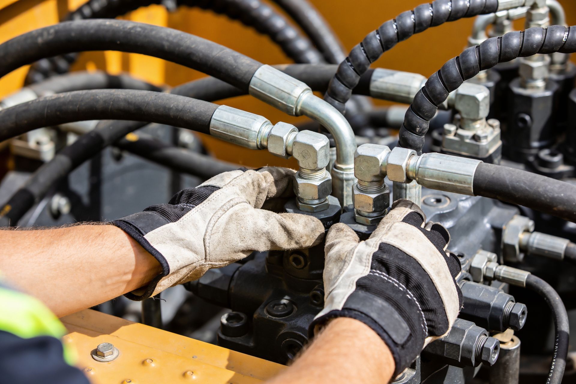 Gloved hands tightening hydraulic hose fittings on heavy machinery.