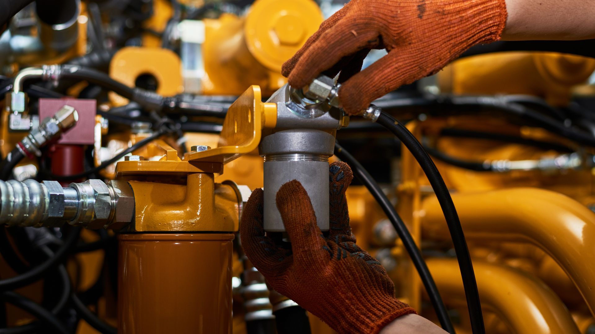 Worker tightening hydraulic fittings on heavy machinery while wearing protective gloves.