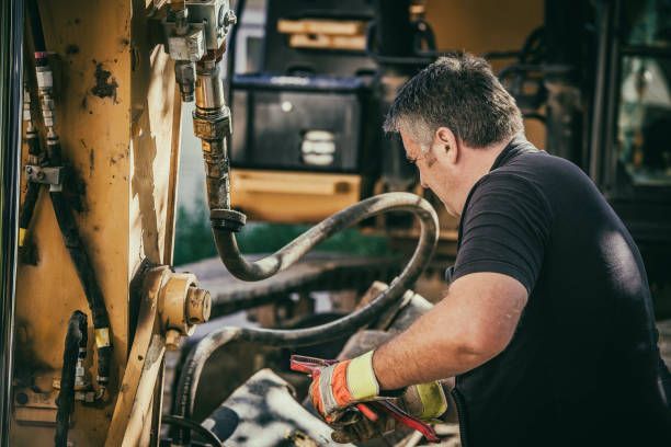 Man in black shirt repairing heavy machinery. Yellow and metallic components, work gloves.