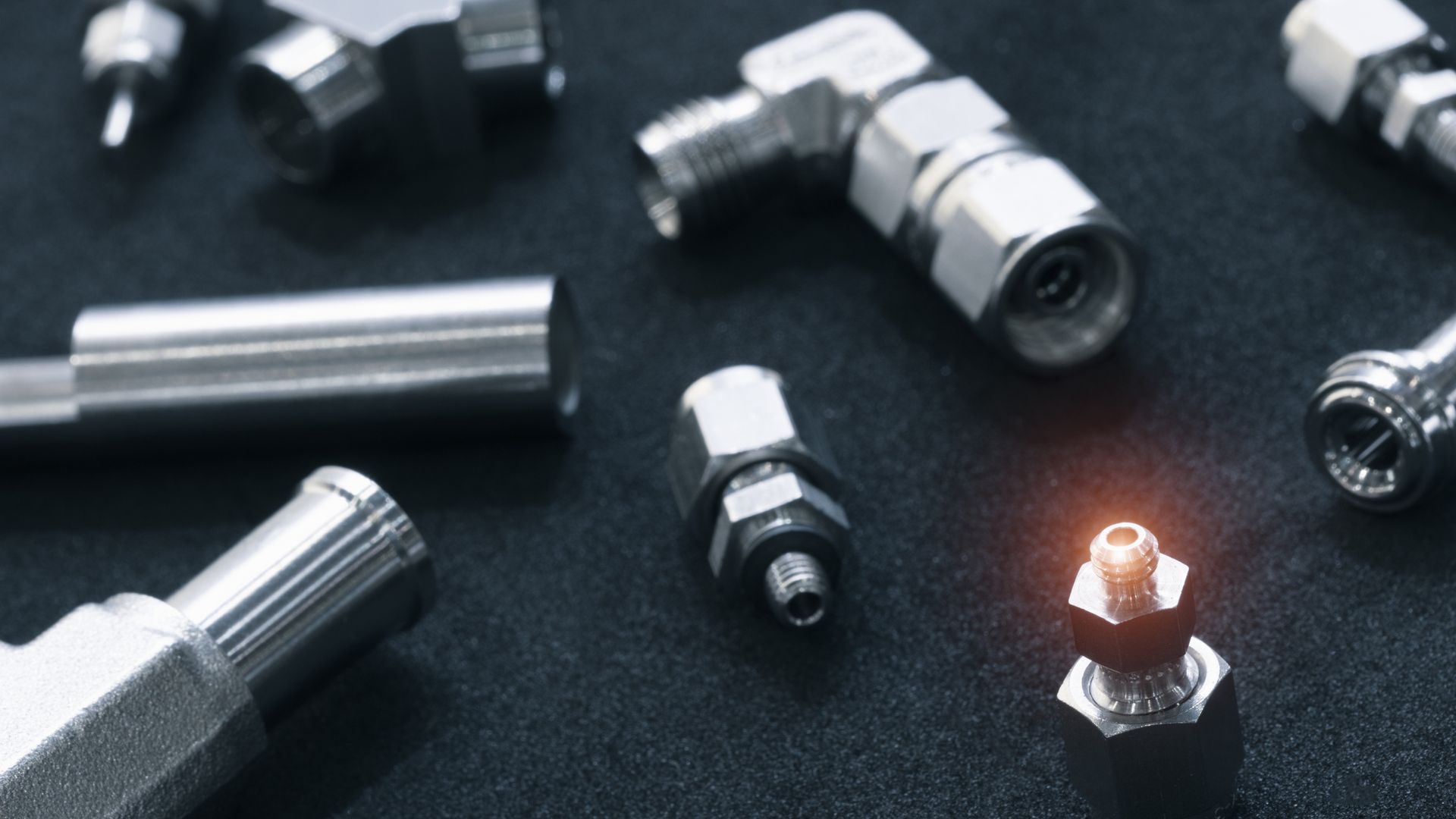 Close-up of various shiny, metallic pipe fittings scattered on a dark surface, some angled, others straight.