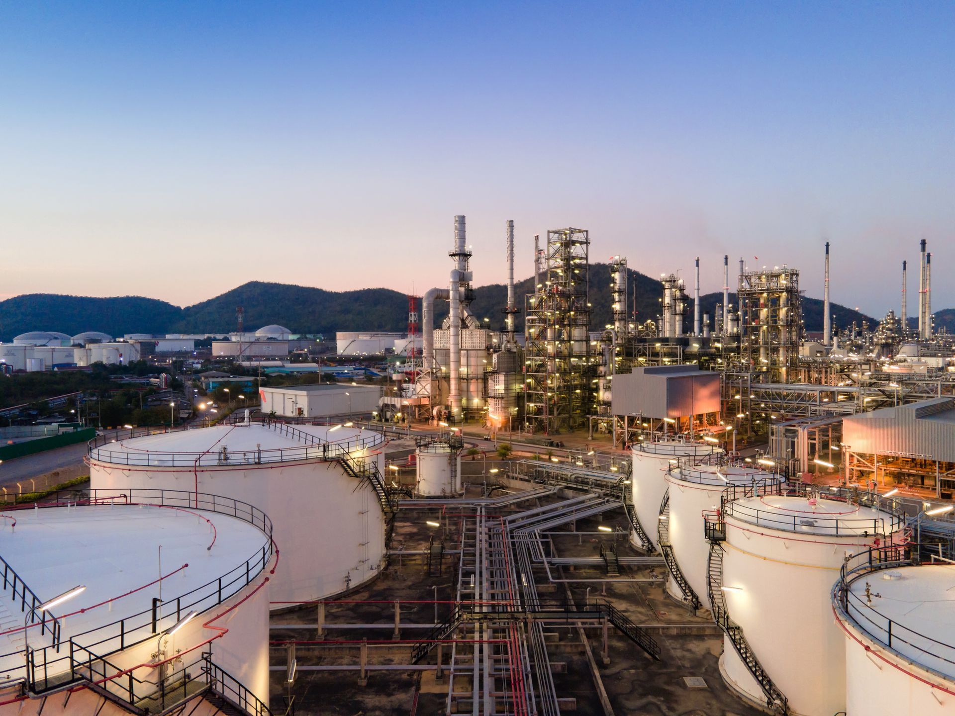 An aerial view of a lit oil refinery complex with storage tanks at dusk. Structures are visible.