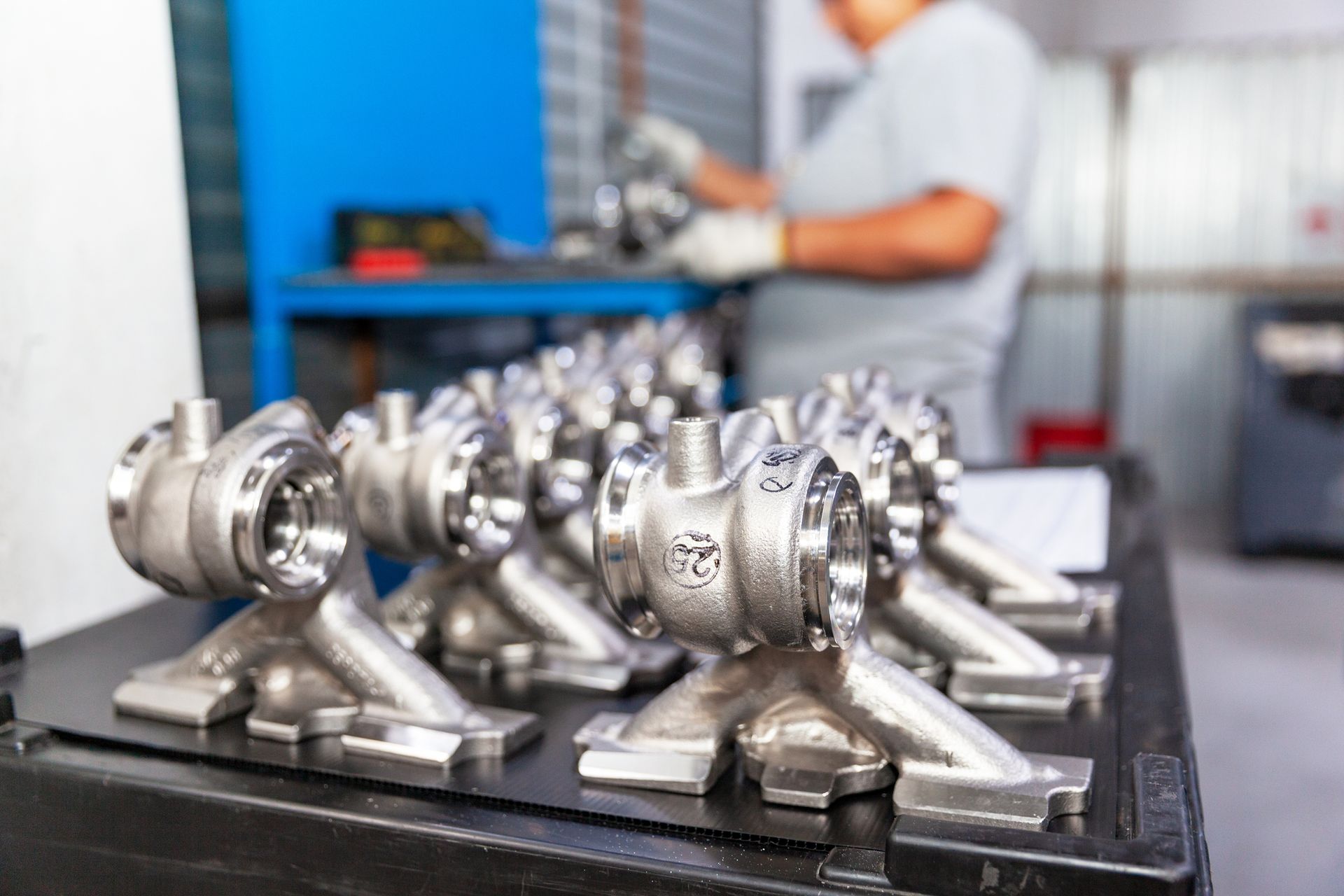 Rows of automotive parts are on top of a table, with a person handling a machine in the background. Rows of automotive parts are on top of a table, with a person handling a machine in the background.