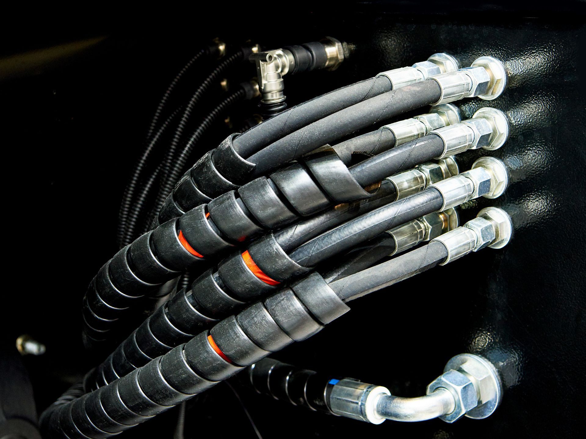 Black hydraulic hoses with protective spiral wrap, connected to fittings on a dark surface.
