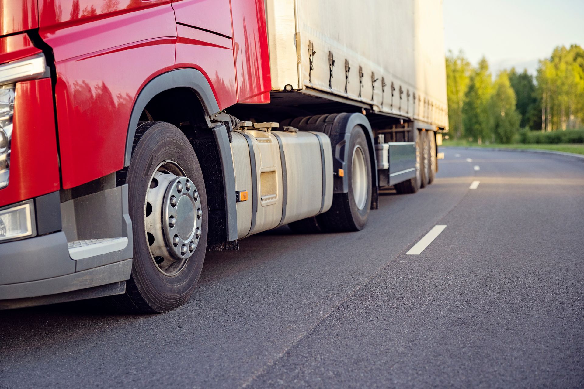 Red semi-truck driving on a paved road with a white line, trees in the background.