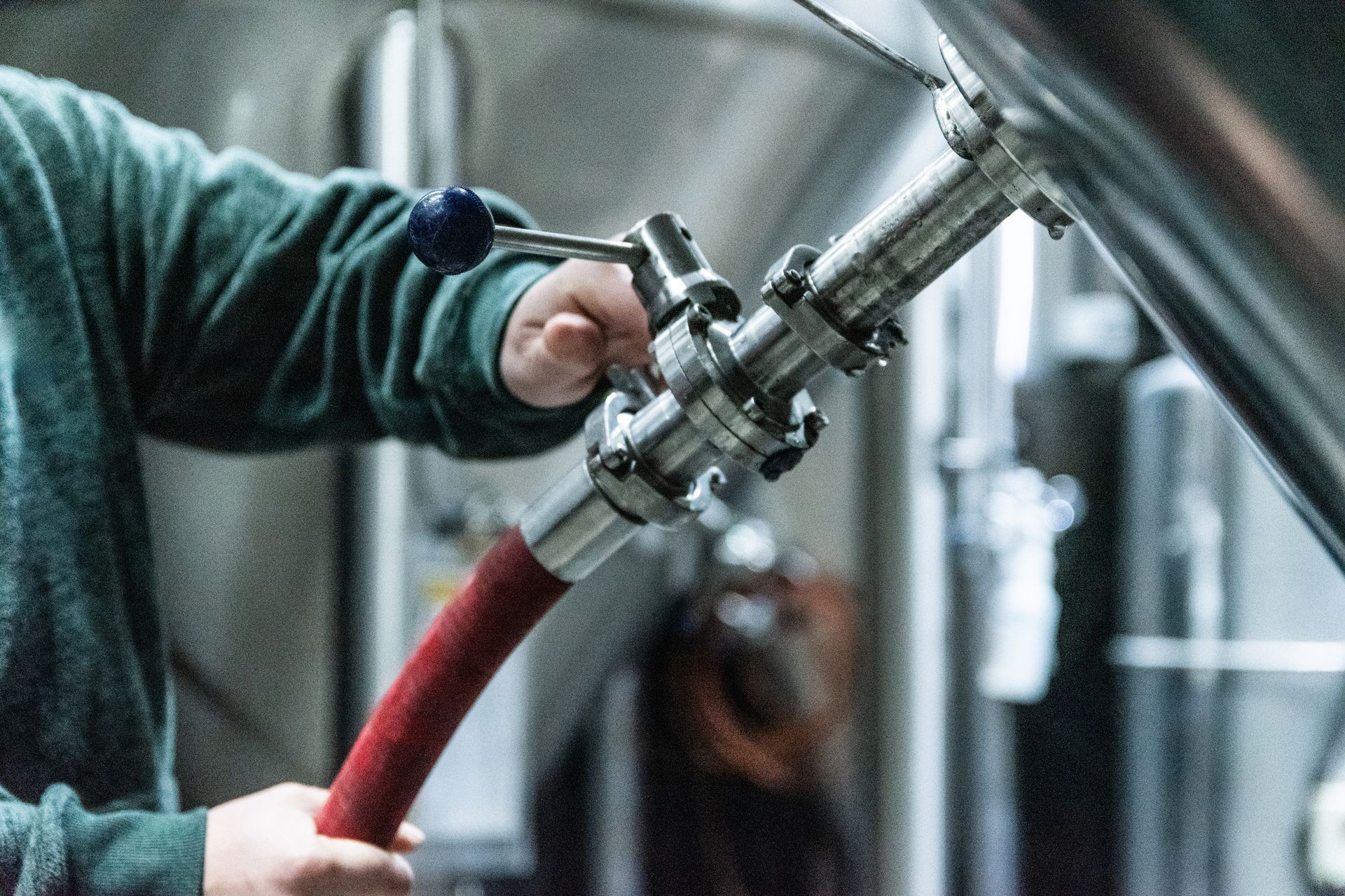 Person in a green sweatshirt operating a valve connected to a red hose near metal brewing equipment.