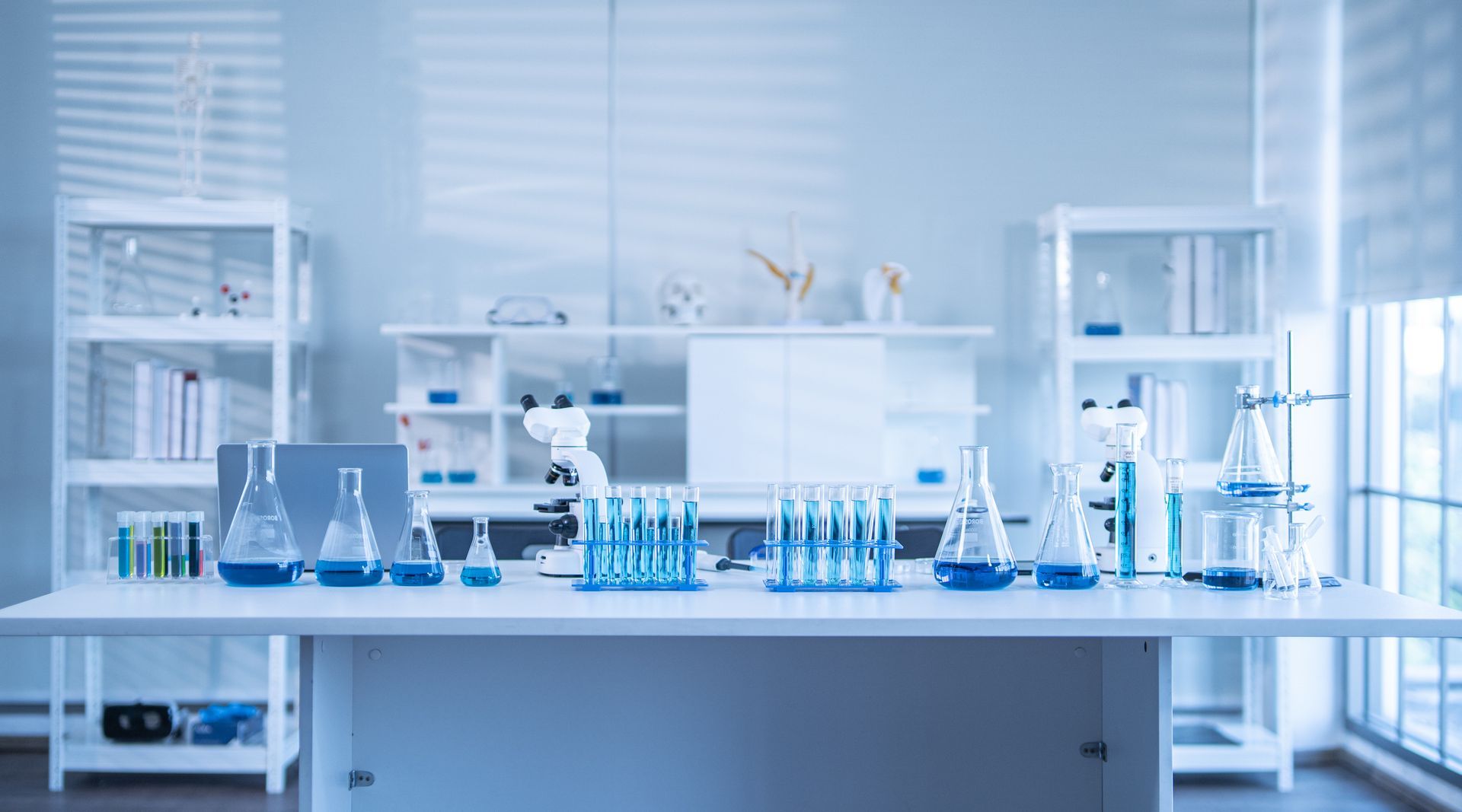 A clean laboratory with a white table holding beakers and test tubes with blue liquid. Shelves hold more lab equipment.