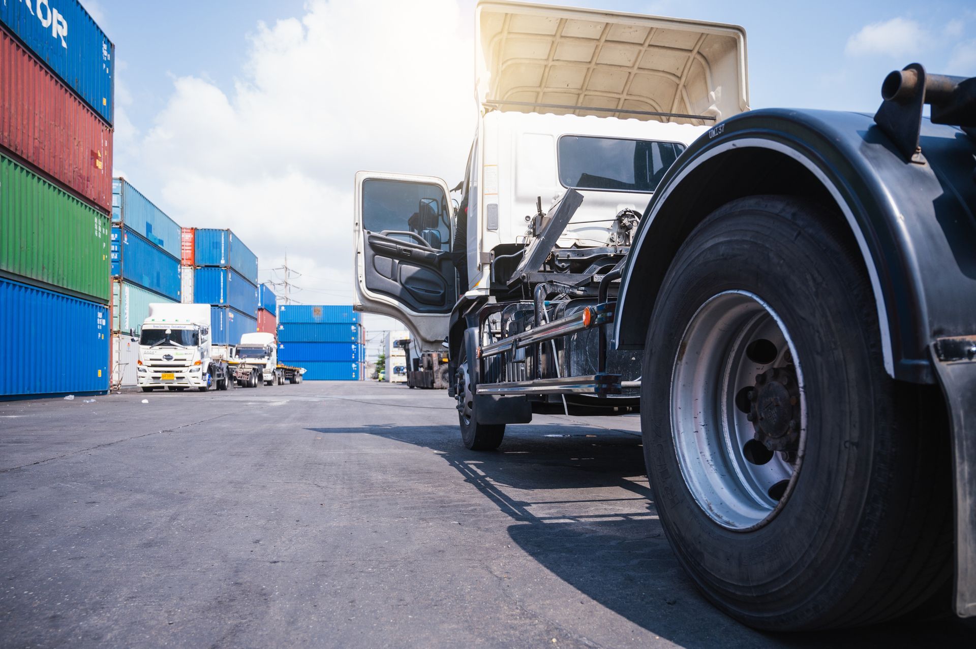 Semi-truck parked in a shipping yard with cargo containers; the truck's door is open, with a bright sun in the background.