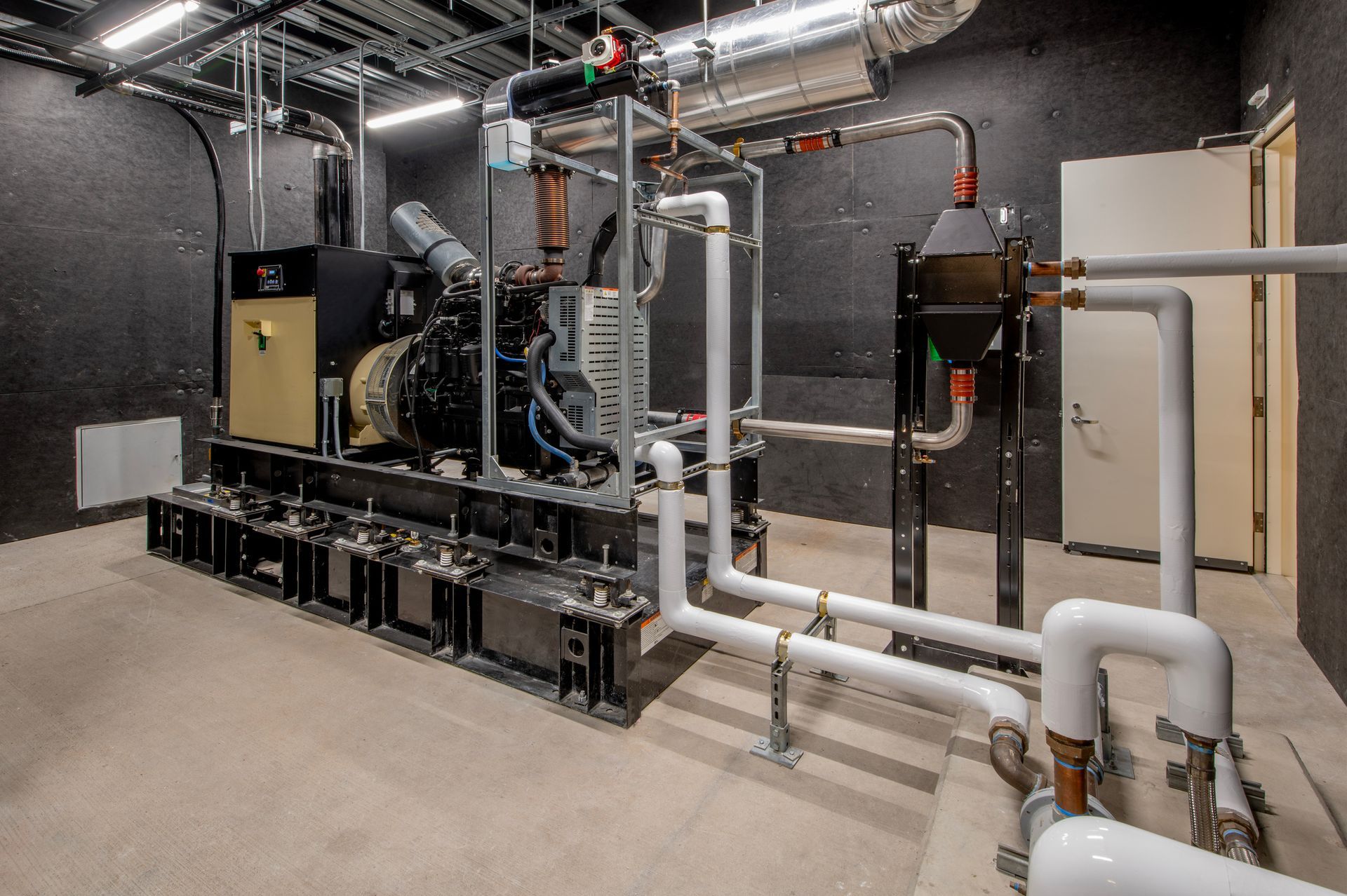 A generator room featuring a large black and gold generator with white pipes and ductwork against dark walls.