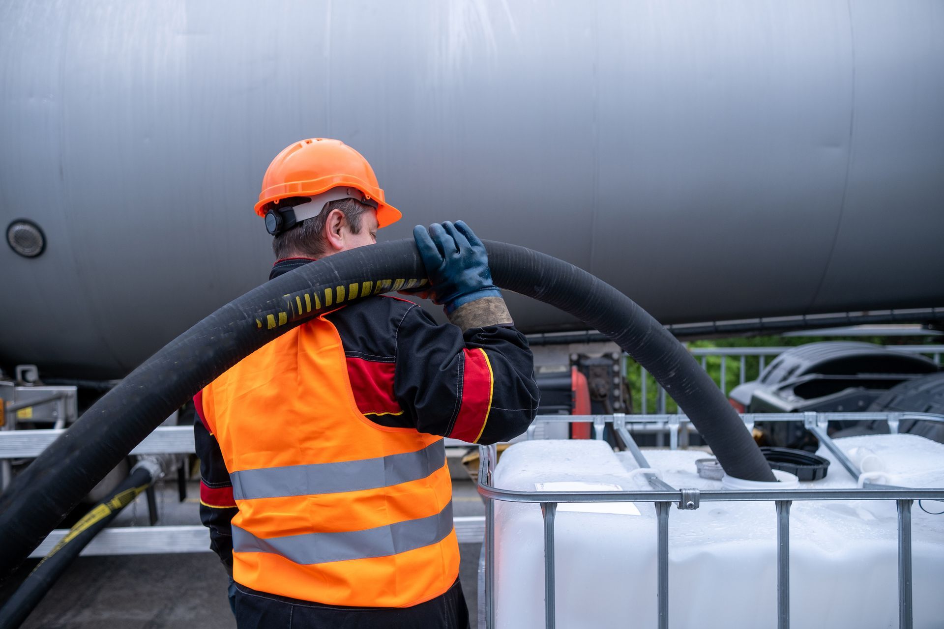 A worker in an orange vest and helmet handles a large black hose, likely for transferring liquid, near a tank and containers.