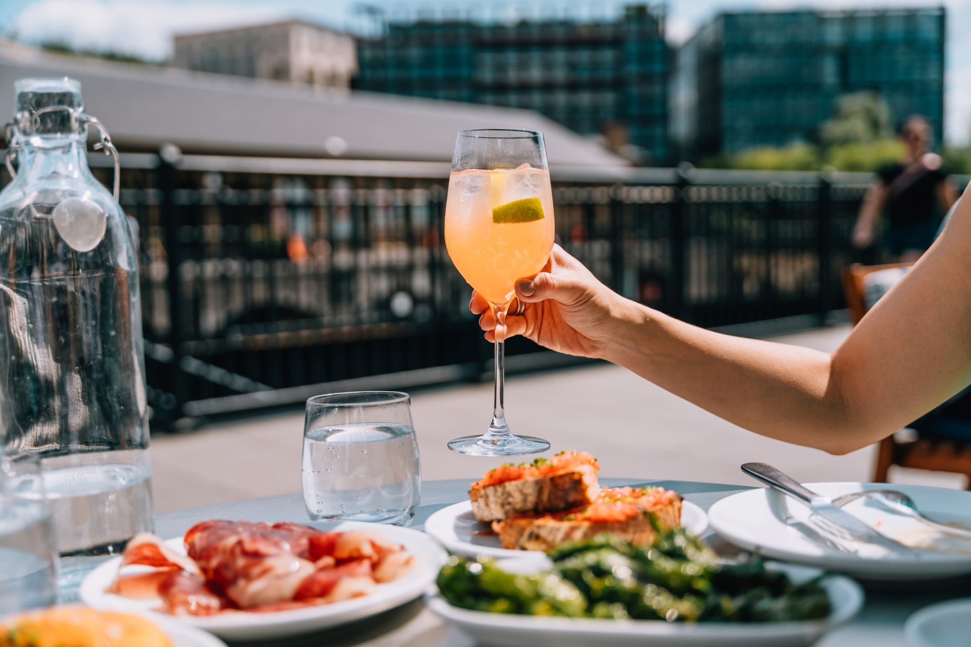 A person holding a mimosa on a rooftop, with food and water bottles on the table. City buildings in the background.