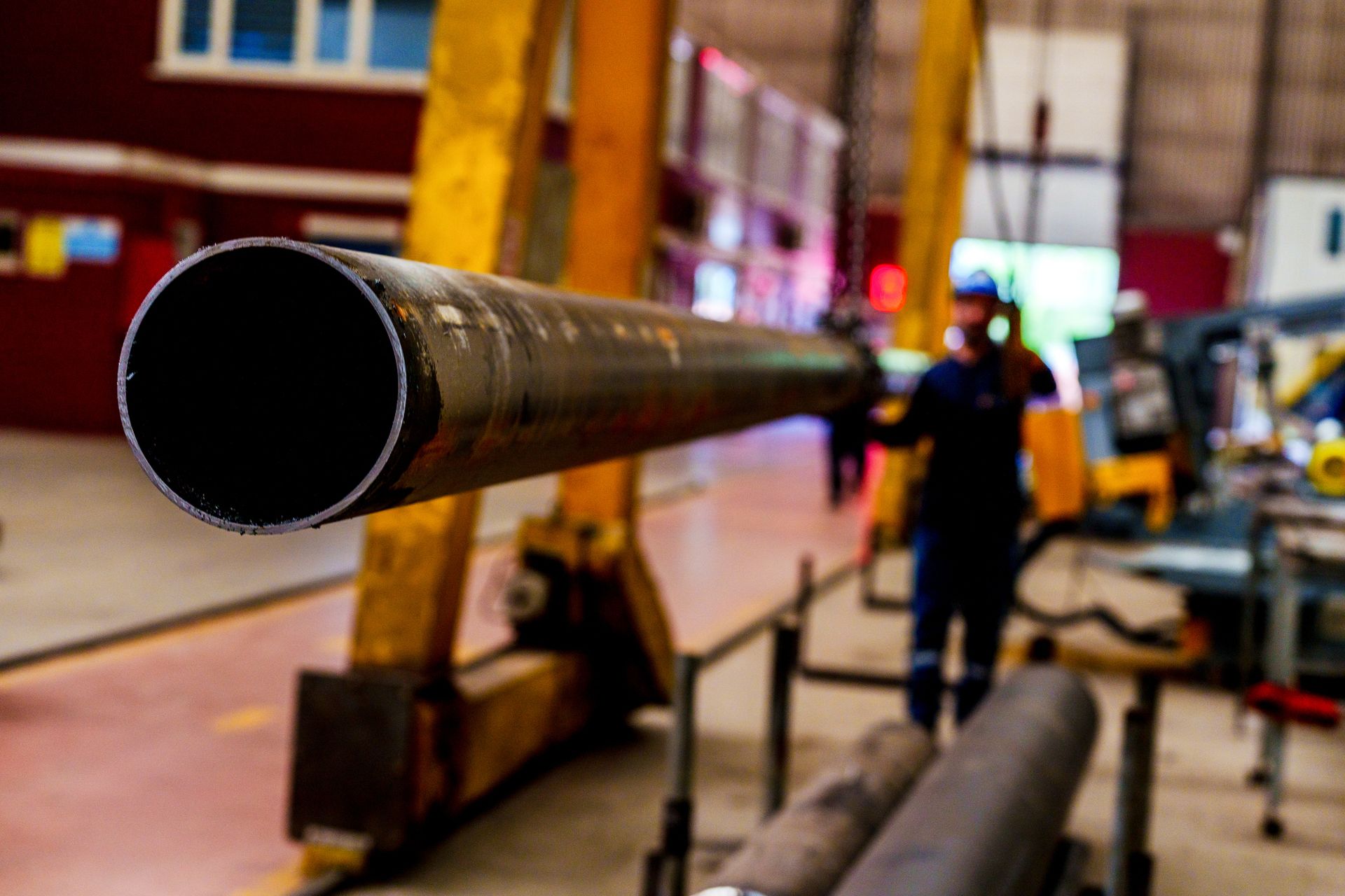 A worker in a blue uniform is lifting a long metal pipe in a factory, supported by a yellow crane.