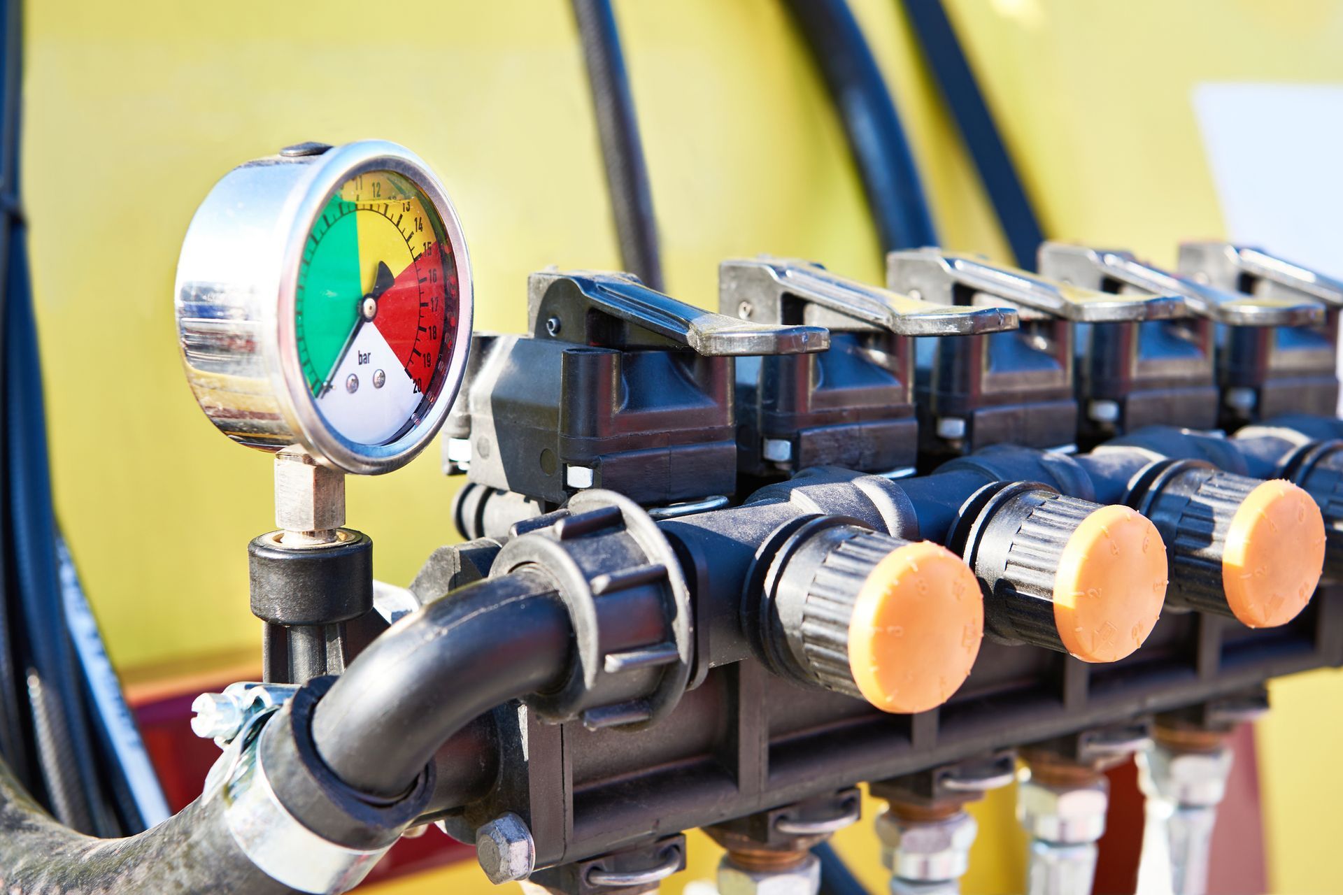 A close-up of agricultural equipment showing a pressure gauge and valves. The gauge has green, yellow, and red sections, and the valves have orange caps.