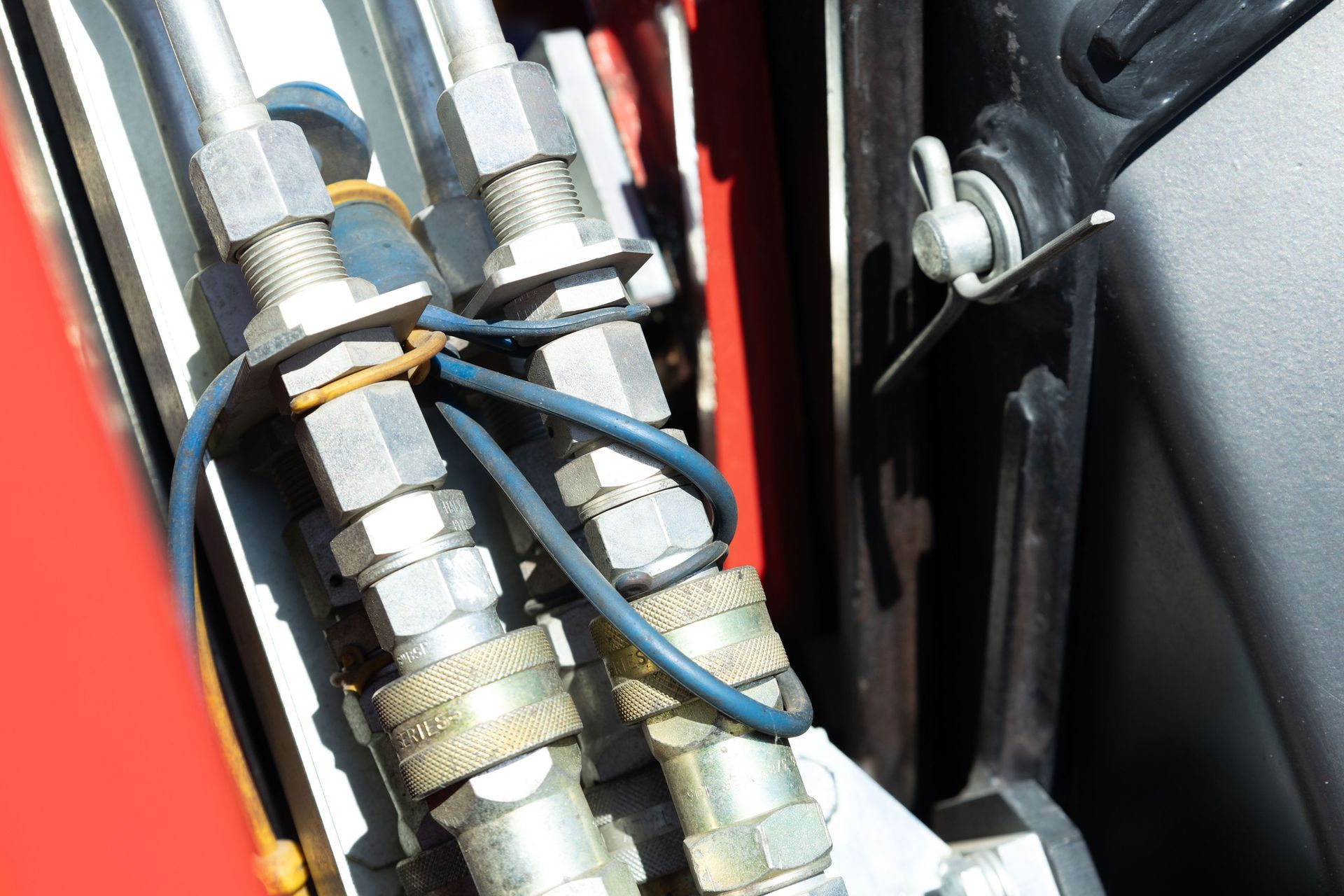 Hydraulic fittings with blue wire and rubber band securing lines on red machinery near black trim.