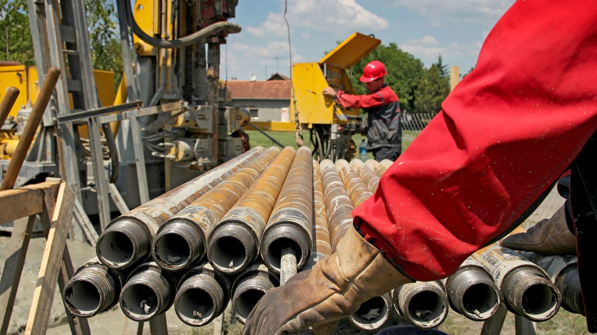 Worker in red jacket and gloves near drilling equipment, rows of metal pipes, sunny outdoor setting.