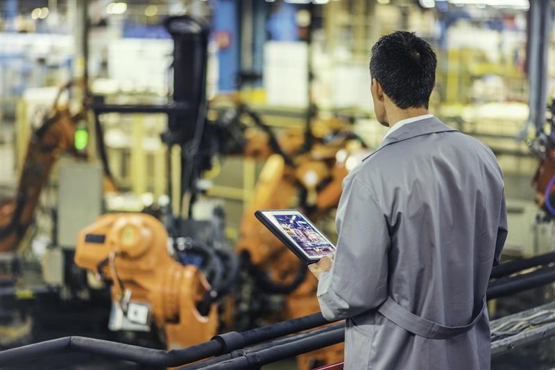 Person in lab coat with tablet observing orange robotic arms in industrial assembly setting.