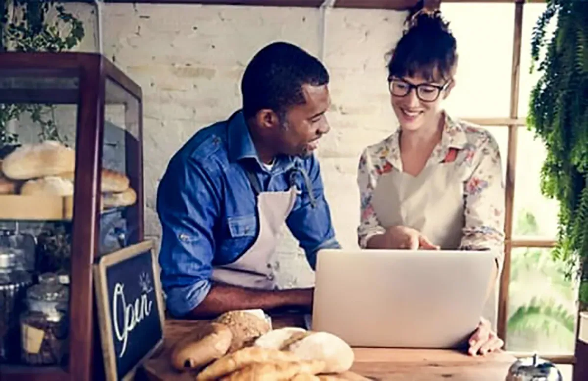 Two people in aprons look at a laptop in a bakery, bread on counter, chalkboard sign 