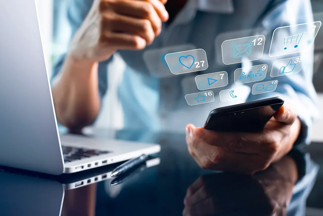 A person using a smartphone and a laptop surrounded by digital social media notification icons in a workspace.