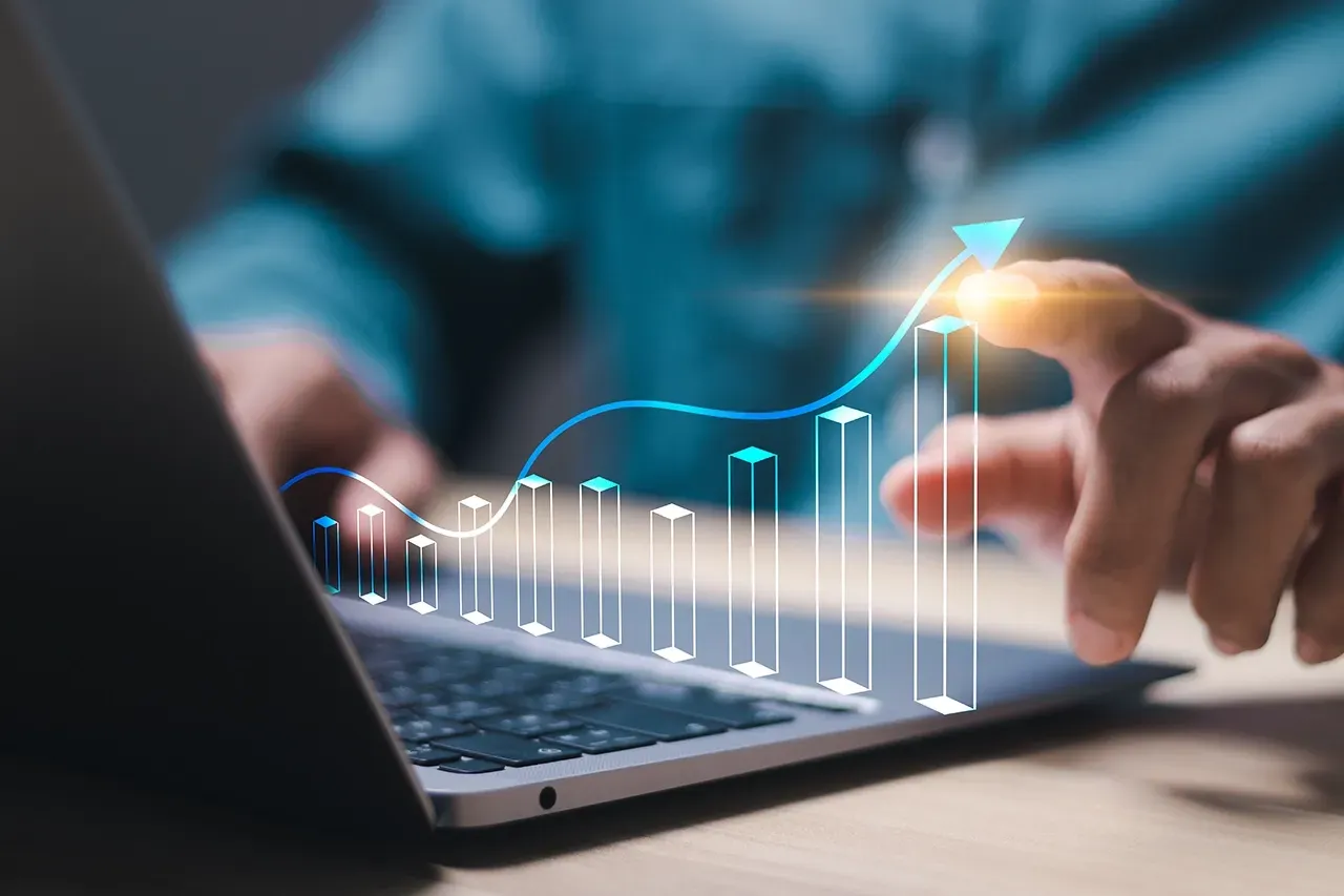 A person interacts with a laptop showing a glowing blue bar chart and upward-trending arrow, symbolizing business growth.
