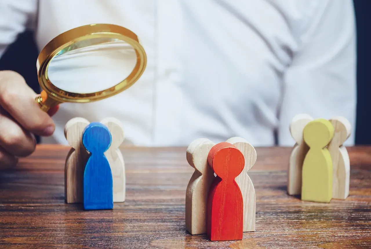 A person holds a magnifying glass over three groups of wooden silhouette figures, each with one figure highlighted in color.