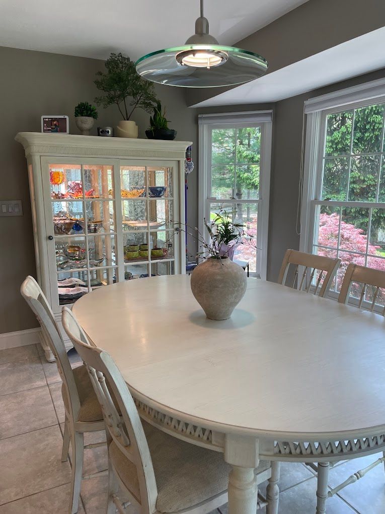 White dining room with oval table, hutch, chairs, and large windows.