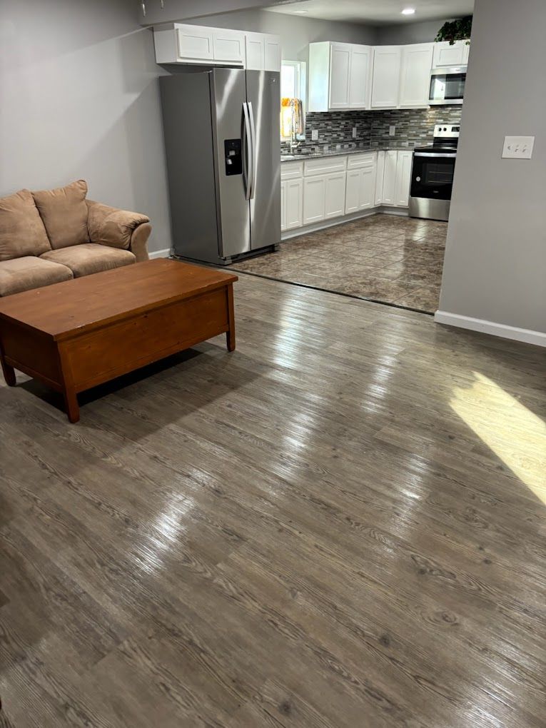 A living room with wood-look flooring, sofa, and a wooden trunk. The kitchen is in the background.
