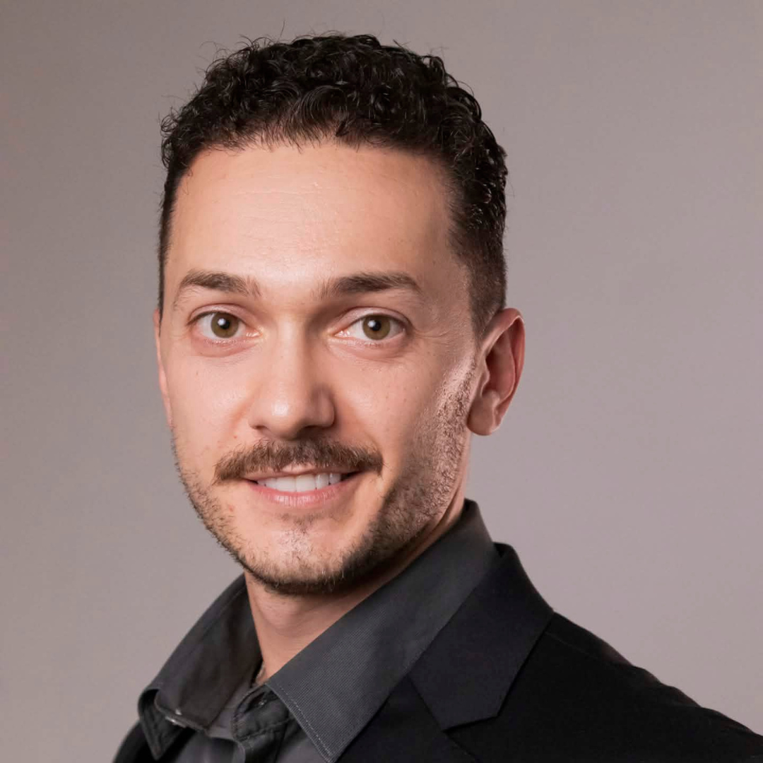 Portrait of a smiling man with short curly hair and a mustache, wearing a dark shirt against a gray background