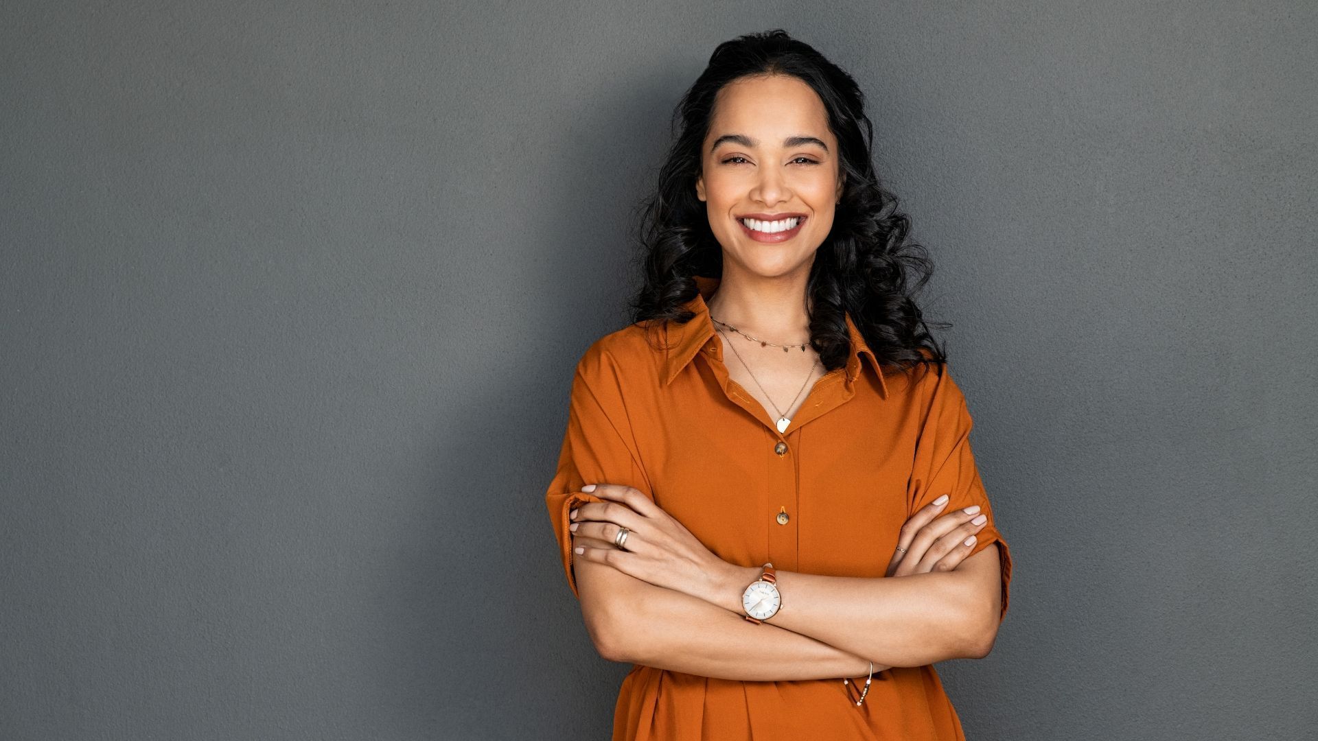 A woman in an orange dress is standing with her arms crossed and smiling.