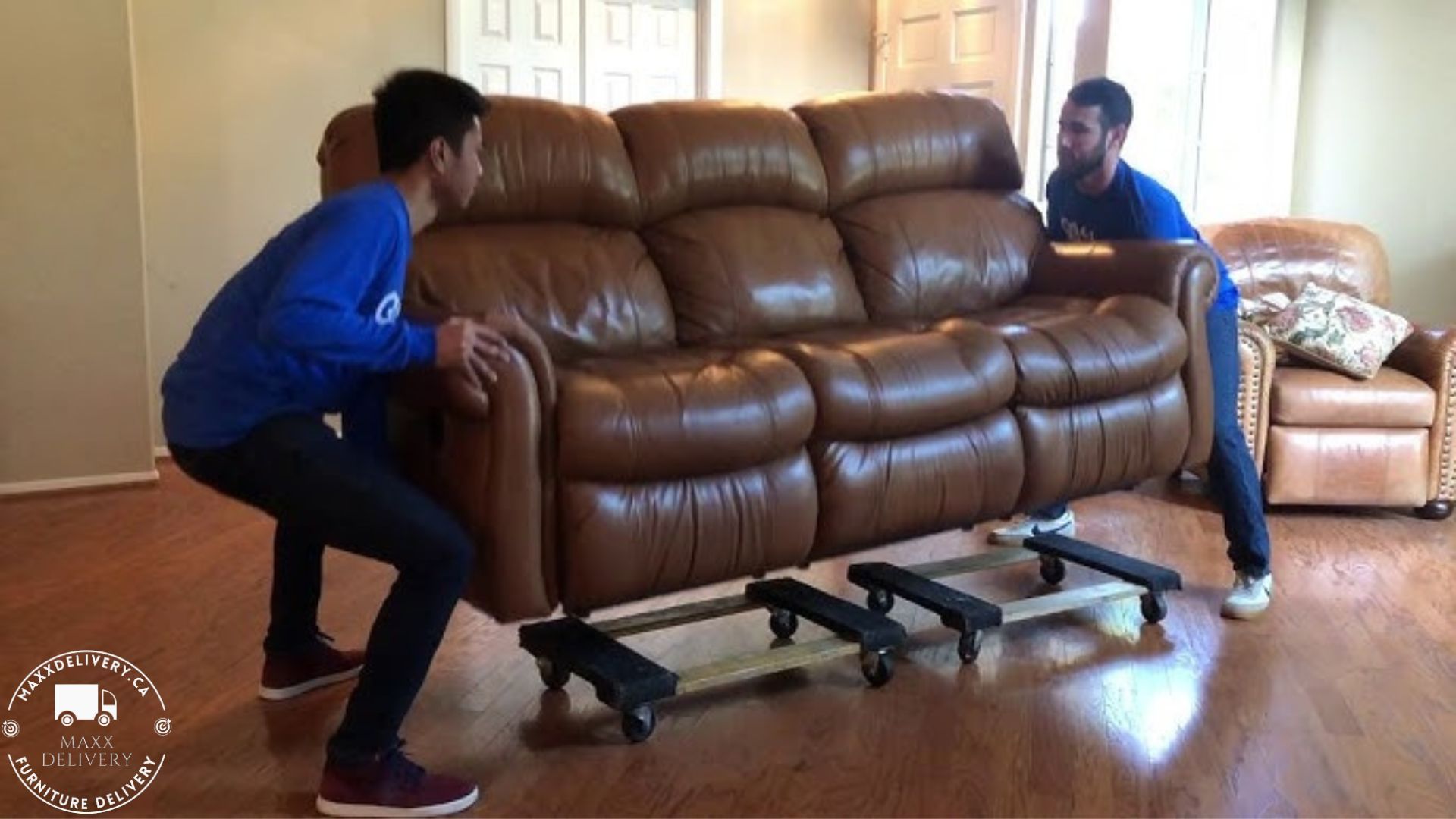 Two men are moving a brown leather couch on wheels