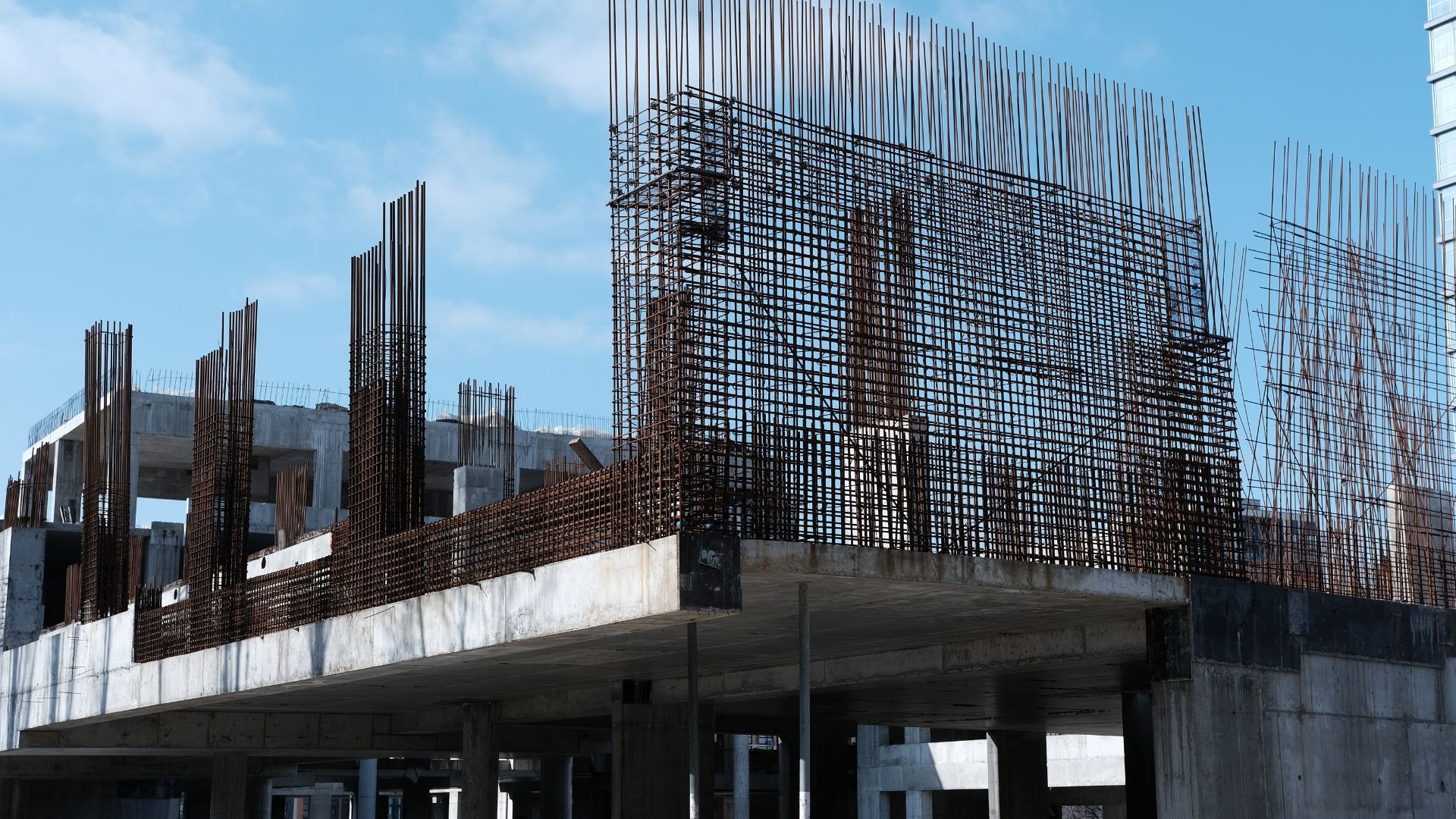 A construction site shows reinforced concrete frameworks with rebar grids against a blue sky.