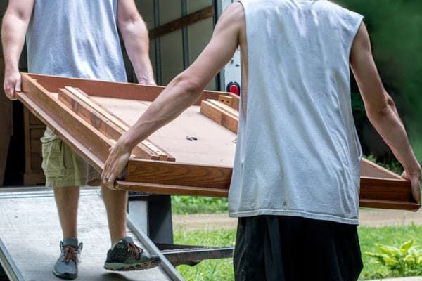 Two men are carrying a wooden table on a truck ramp.