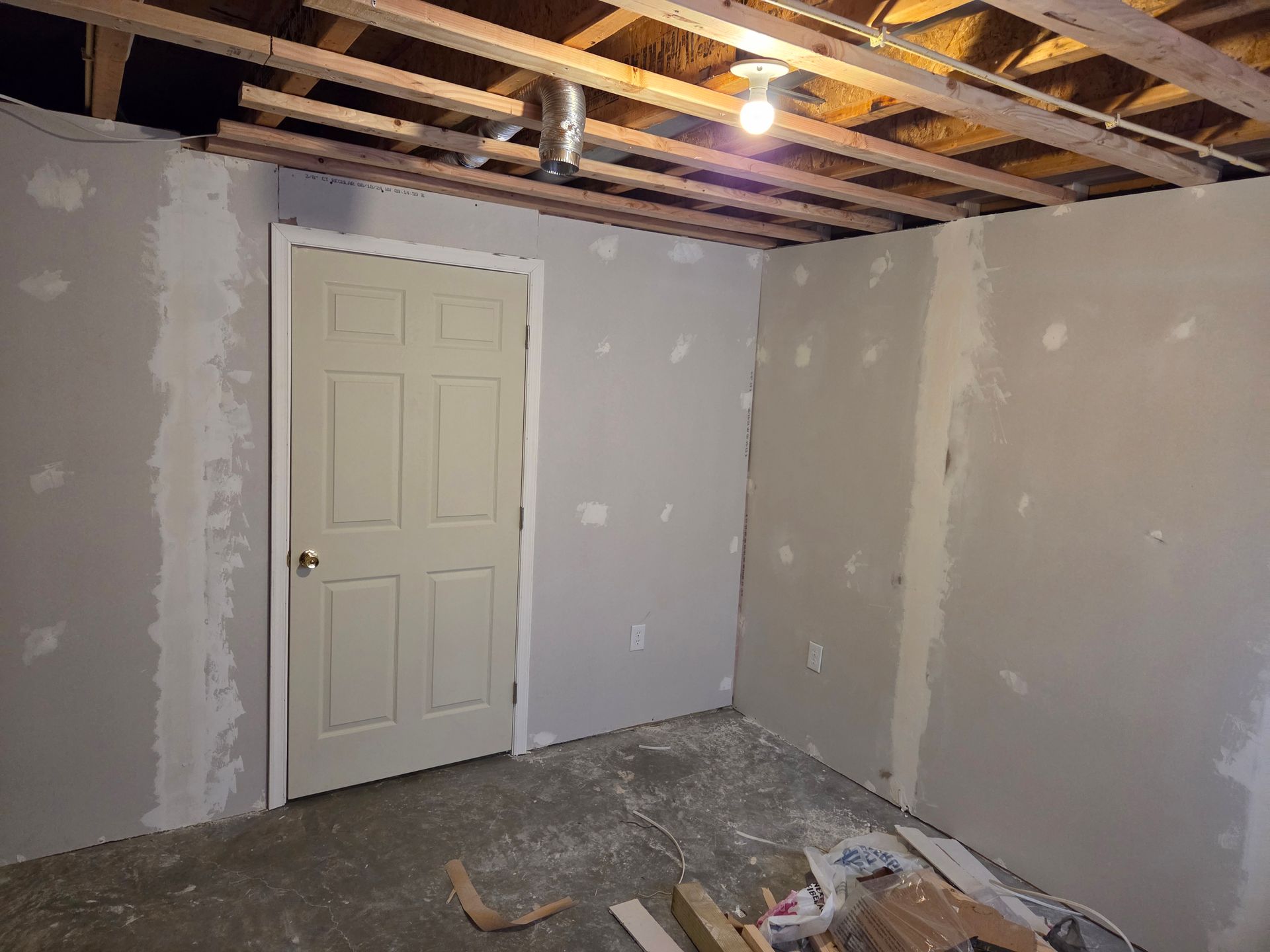 An unfinished basement room with grey drywall, an off-white door, exposed wooden ceiling joists, and a concrete floor.