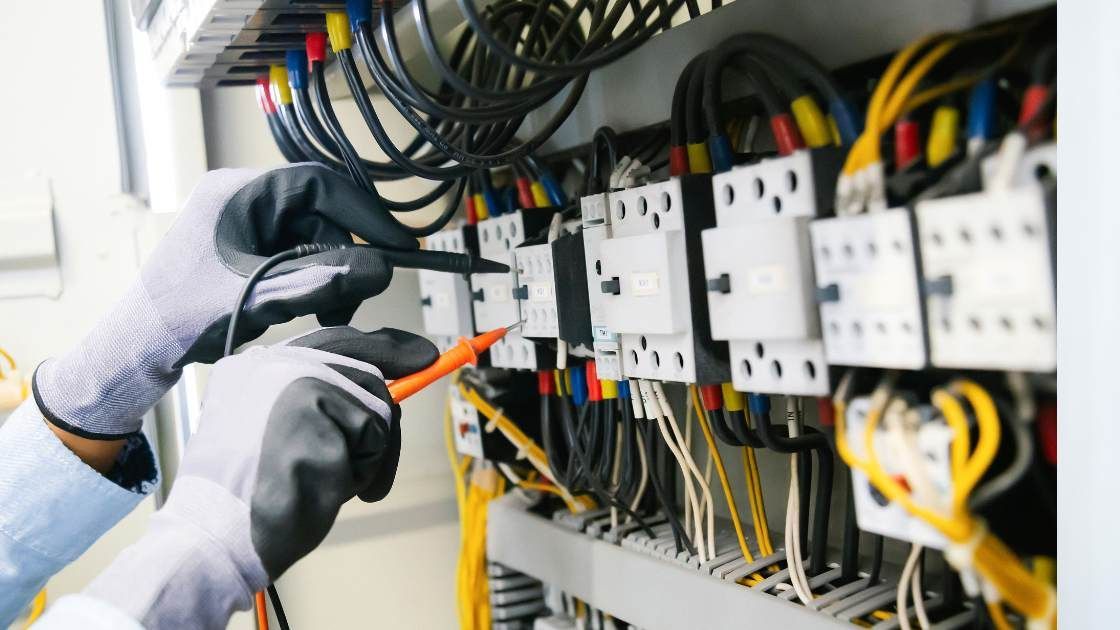 An electrician wearing protective gloves uses a multimeter probe to test electrical components in a control panel.