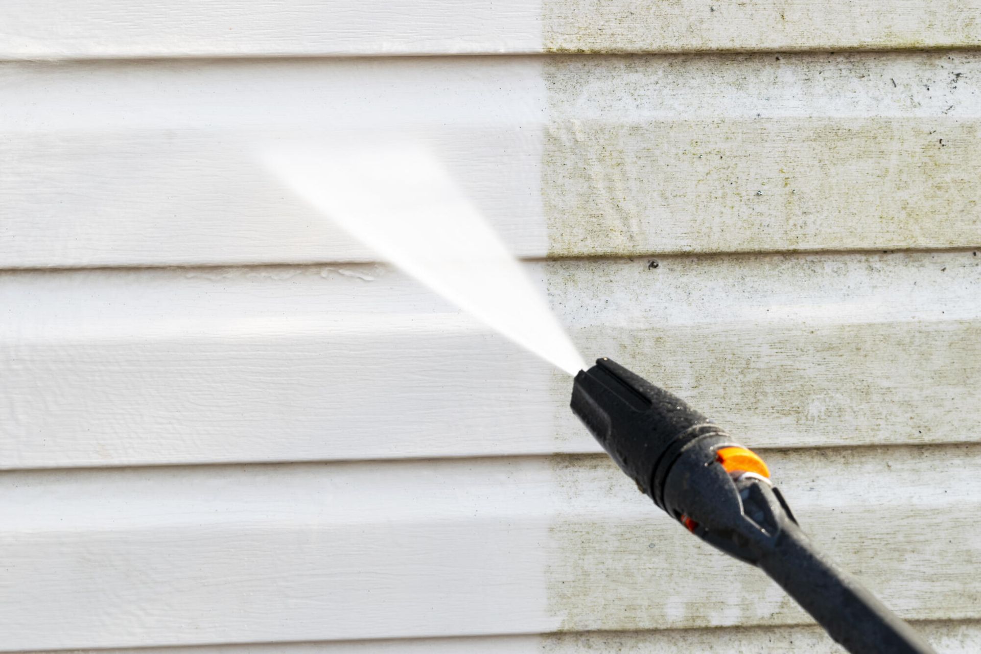 A pressure washer nozzle sprays a stream of water, cleaning grime off light-colored horizontal vinyl siding.
