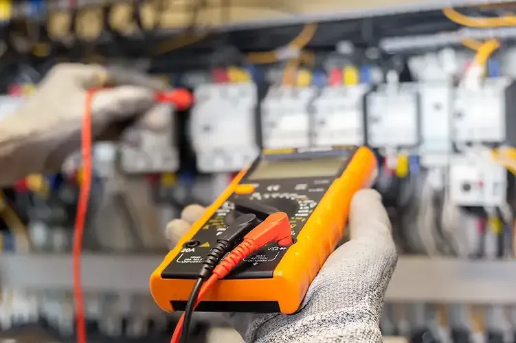 A gloved hand holds a yellow multimeter while another hand uses a red probe to test electrical components in a panel.