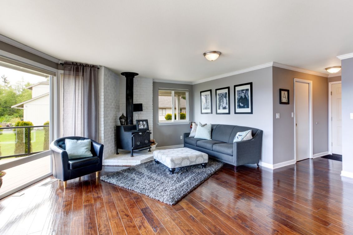 Living room with dark wood floors, a grey sofa, matching armchair, wood-burning stove, and a grey area rug.