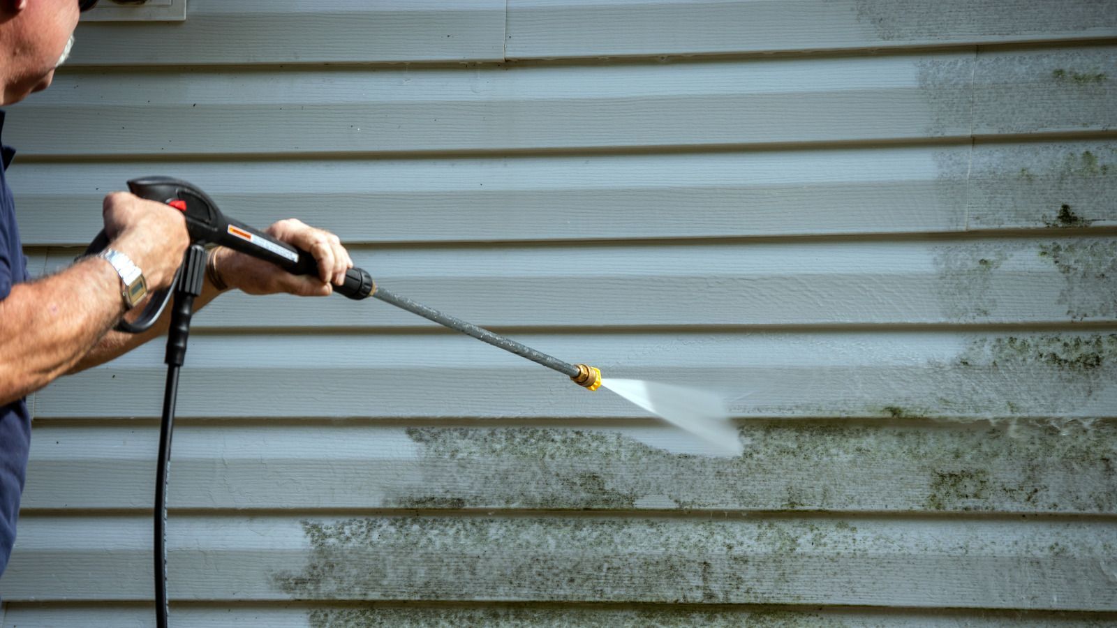 A person pressure washing grime off grey horizontal siding, showing a clean area next to a dirty one.