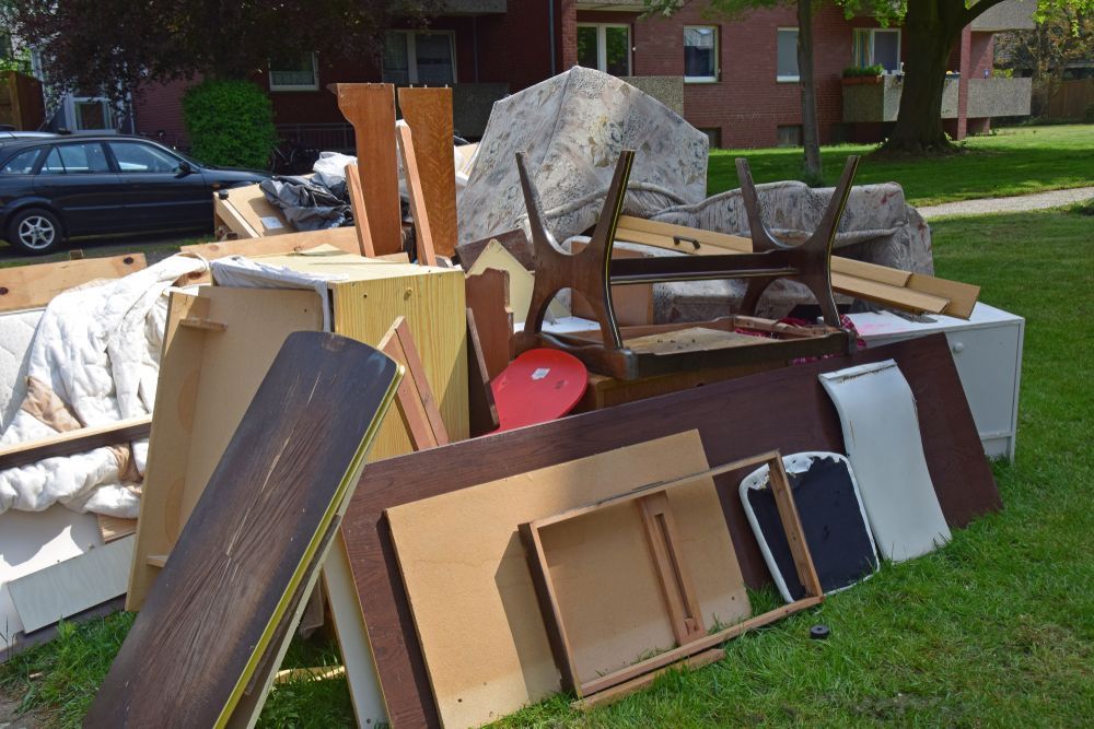 A pile of discarded furniture and household items sits on the grass by a brick building.