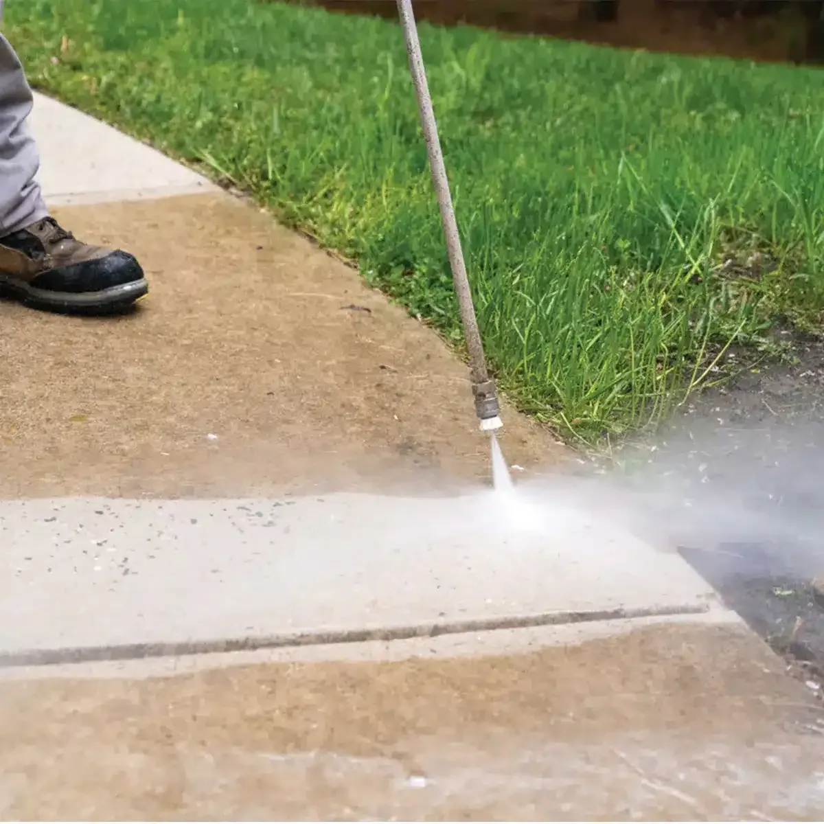 A person power washing a concrete sidewalk next to a green lawn, showing the contrast between dirty and clean surfaces.