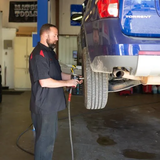Mechanic using a tool on a car wheel in a garage; blue car elevated on a lift.